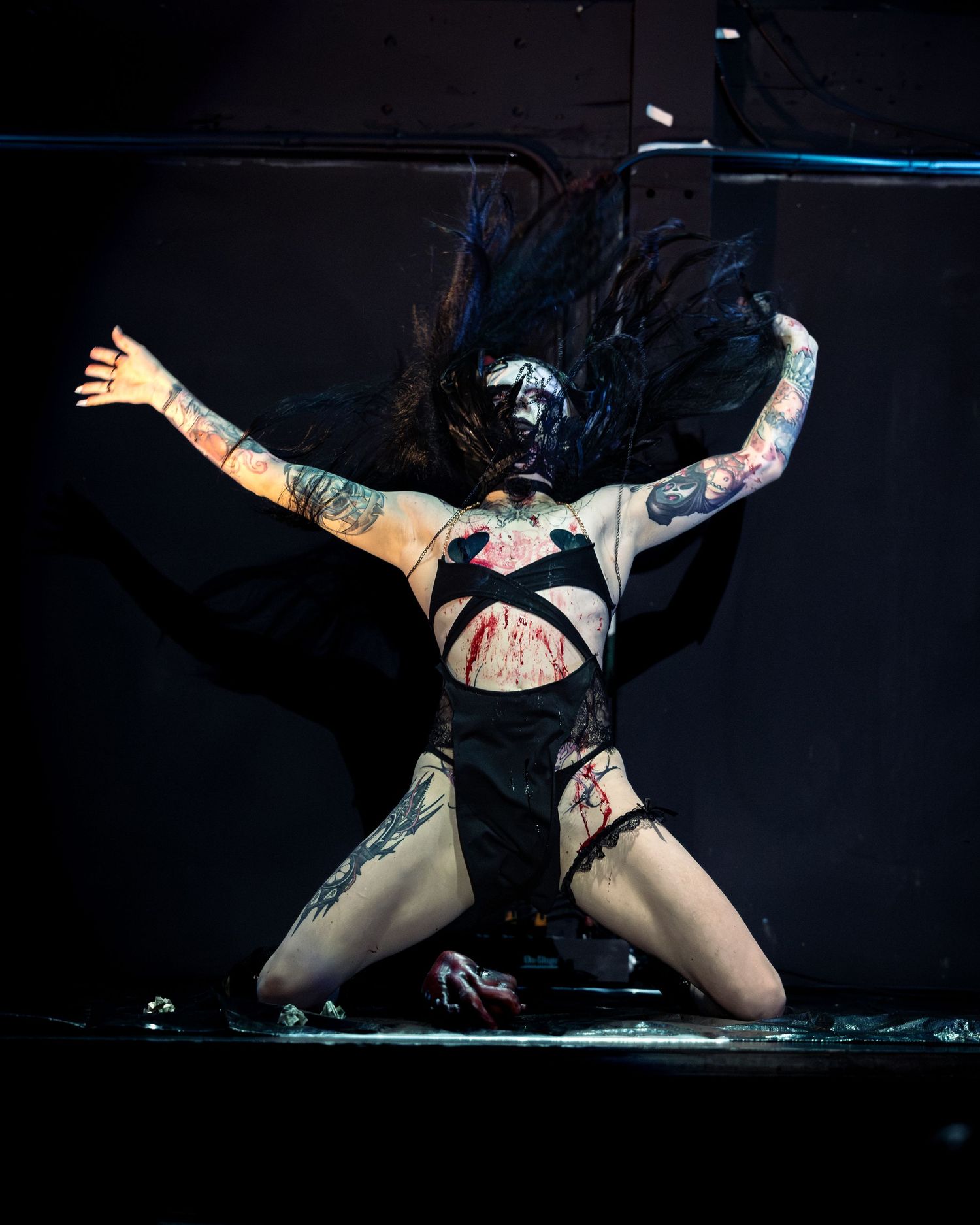 Tattooed performer in dark costume with fake blood kneels dramatically on stage with arms outstretched and hair flying.
