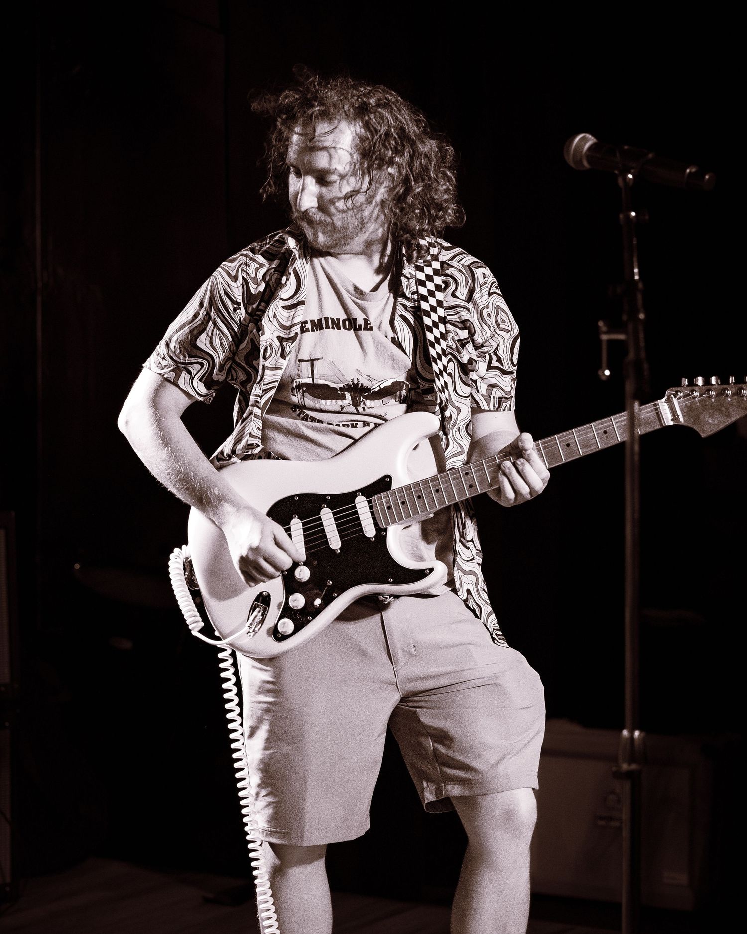 Curly-haired guitarist plays Fender Stratocaster in black and white concert photo.