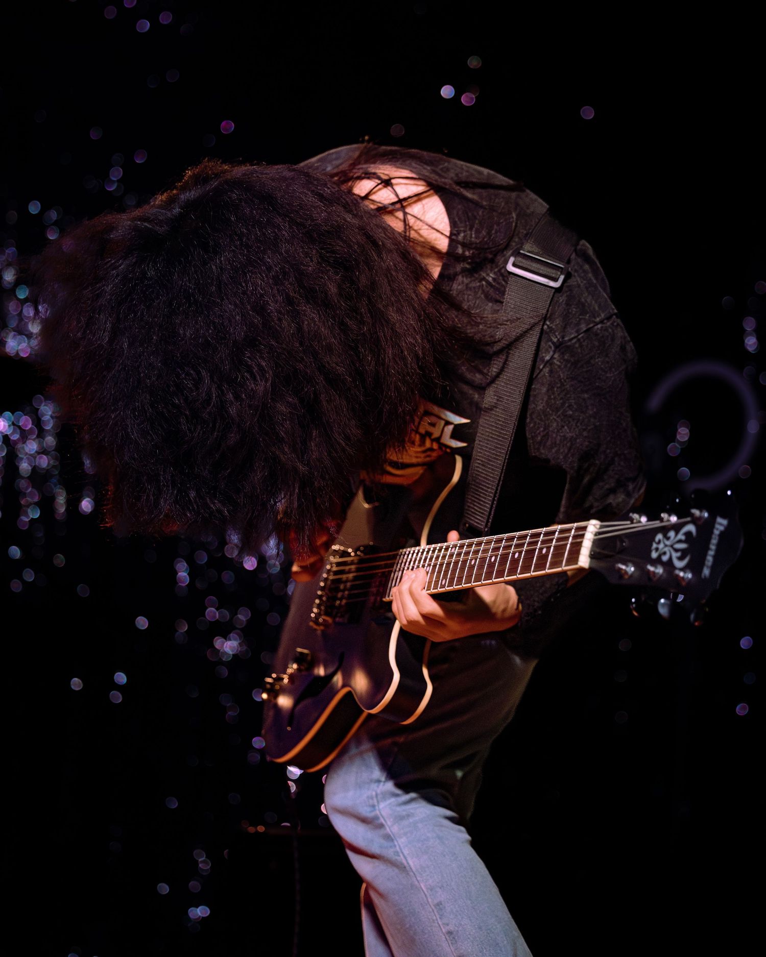 Rock guitarist headbanging with long dark hair covering face while playing electric guitar on stage.