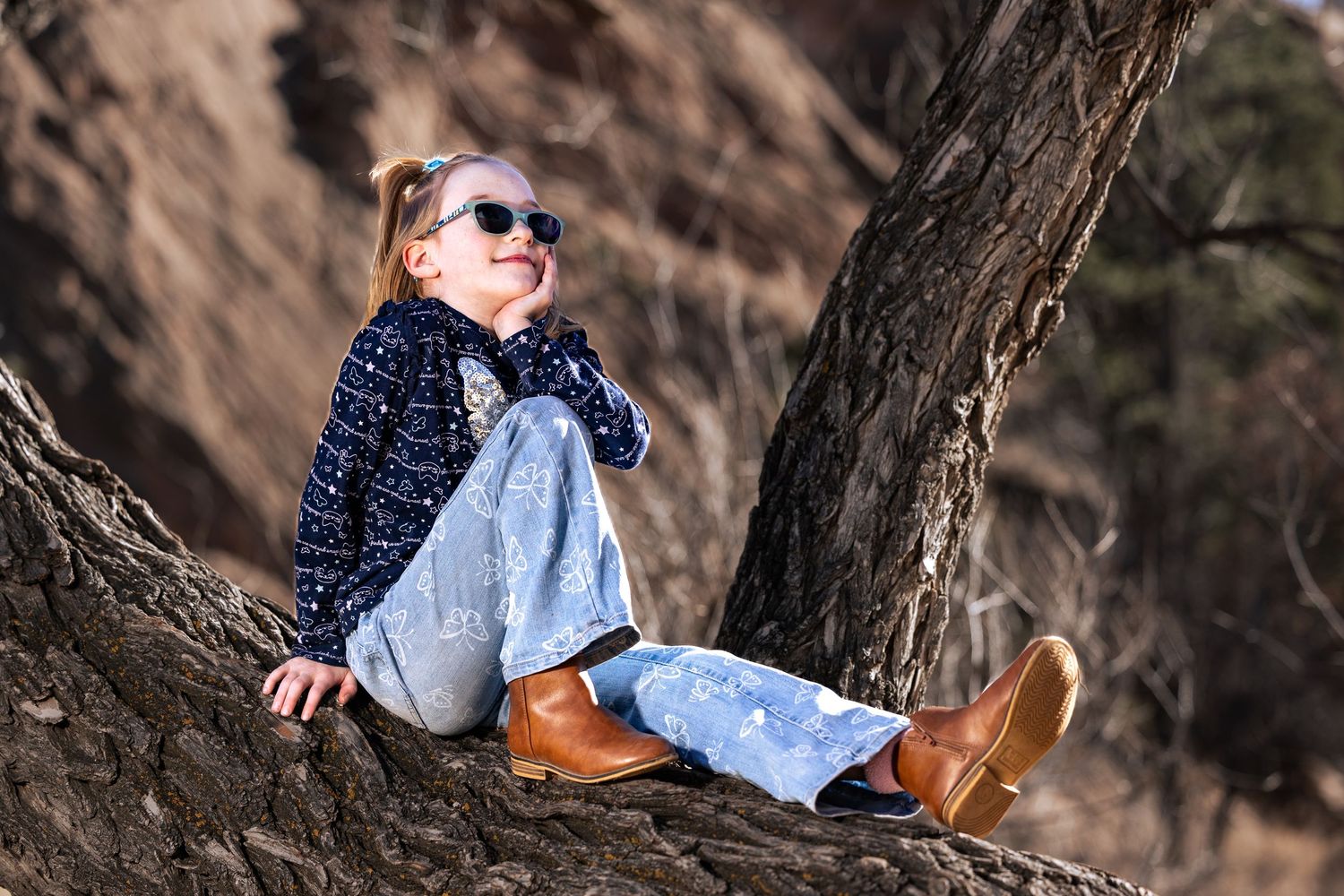 A young girl in sunglasses, jeans, and brown boots sits smiling on a tree trunk in a natural outdoor setting.