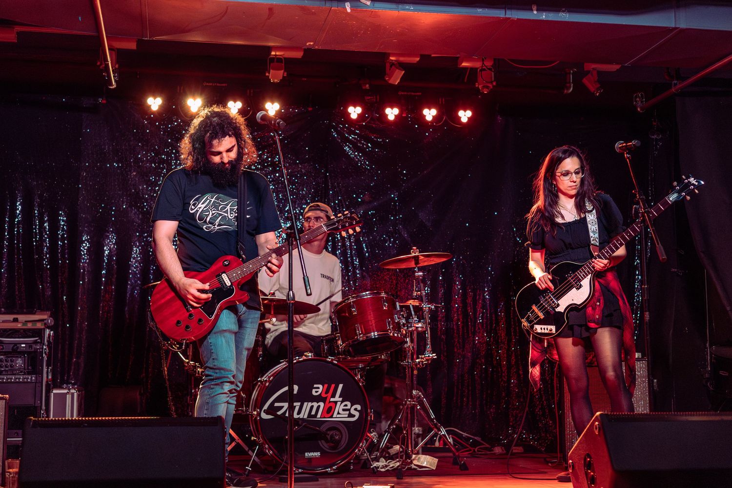 Rock band performing on stage with guitars and drums under red stage lights.