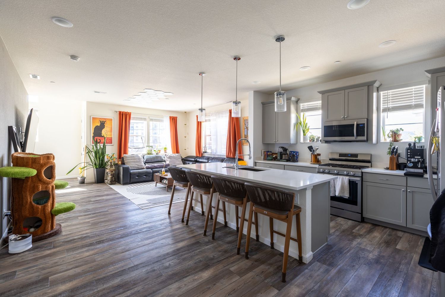 Modern open-concept kitchen with white island, bar stools, gray cabinets, and living room with orange curtains.