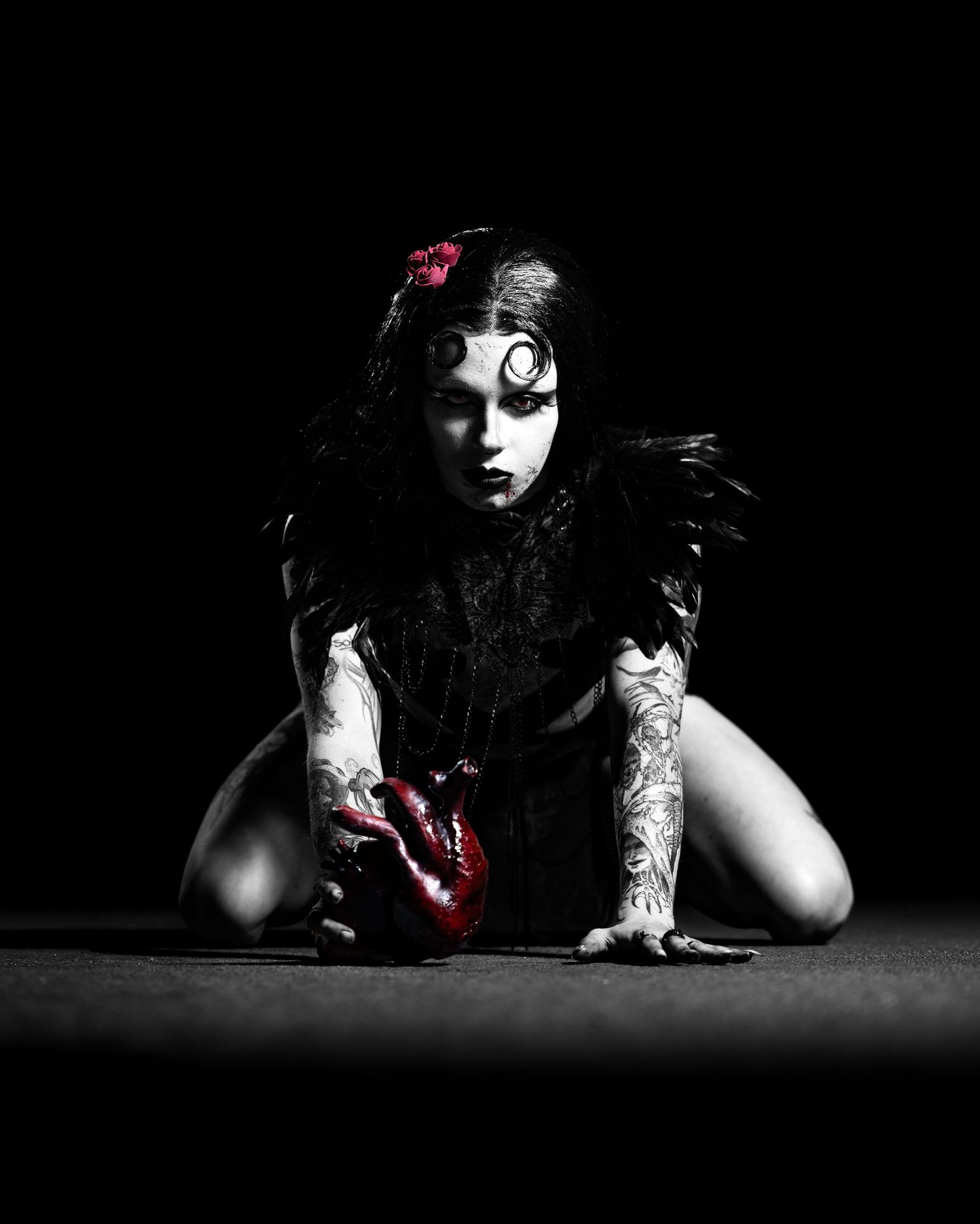 Dark gothic woman in black feathers with tattoos holds a red heart, dramatic black and white artistic photo.