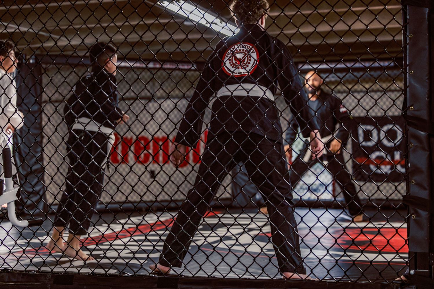 Training session through chain-link fence showing athletes exercising on red mats.