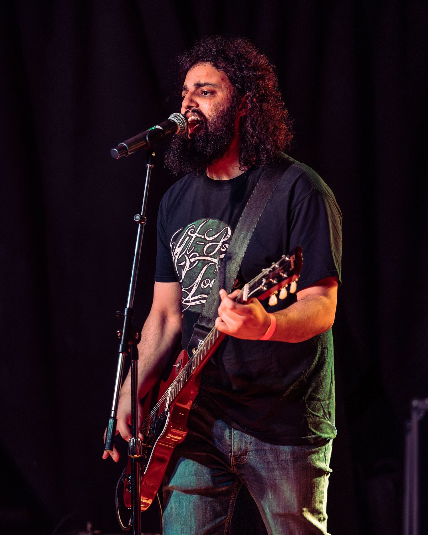 Bearded man with curly hair playing red electric guitar and singing at microphone on stage.