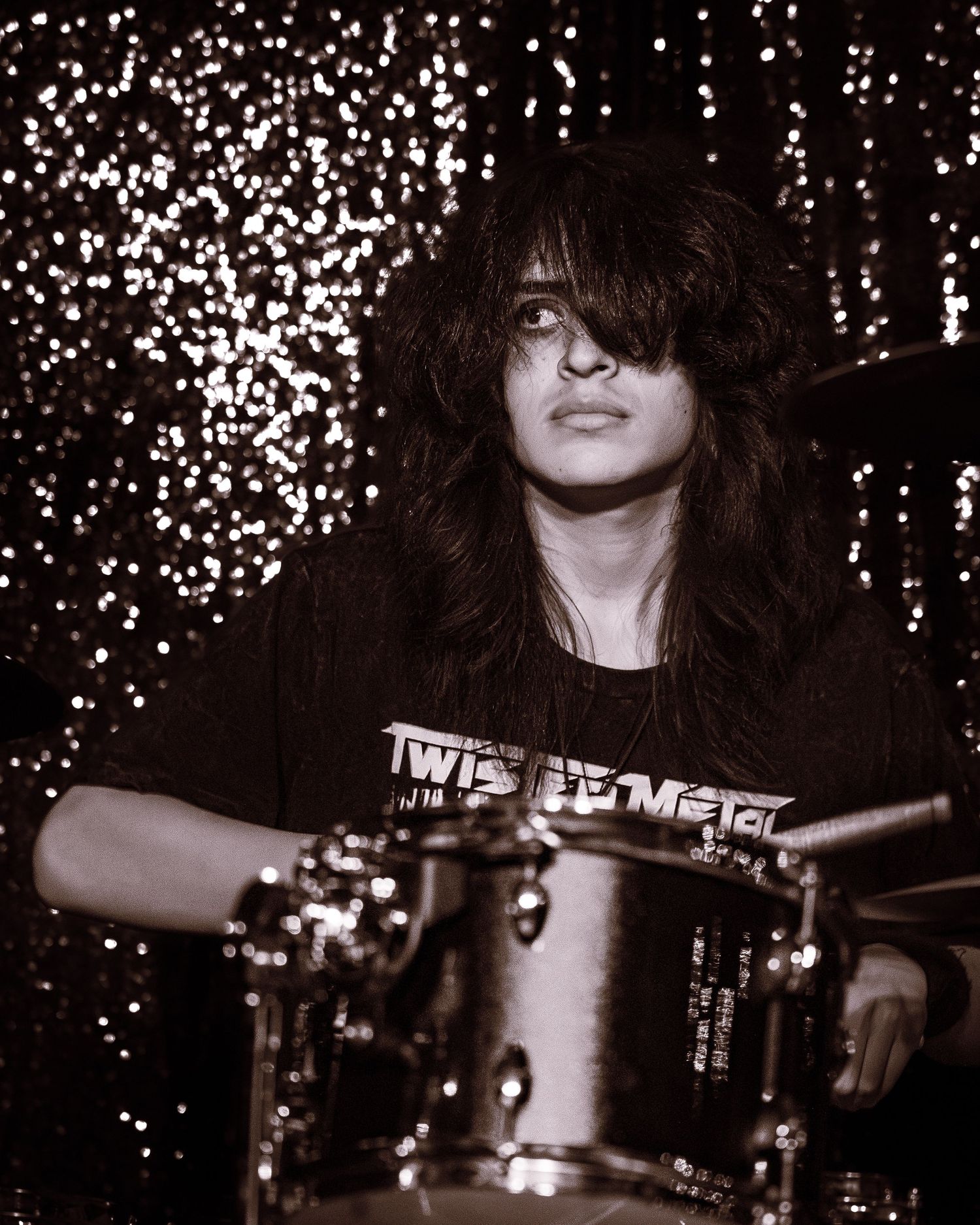 Long-haired drummer playing drums on stage in front of a sparkling backdrop.