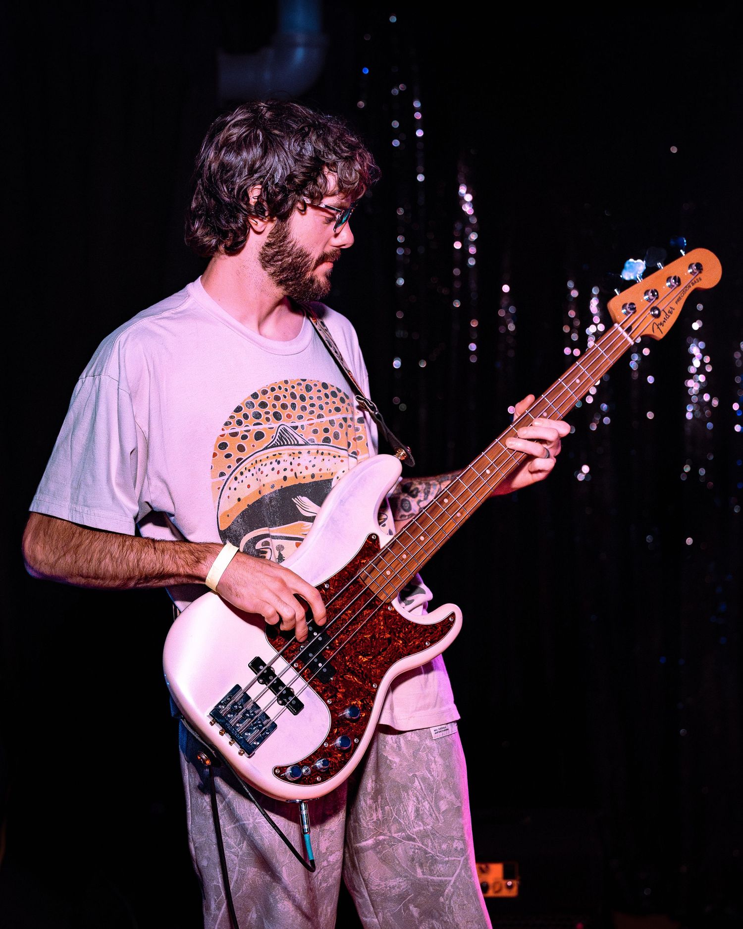 Bearded bassist in white graphic tee plays a pink Fender bass guitar on a dark concert stage.