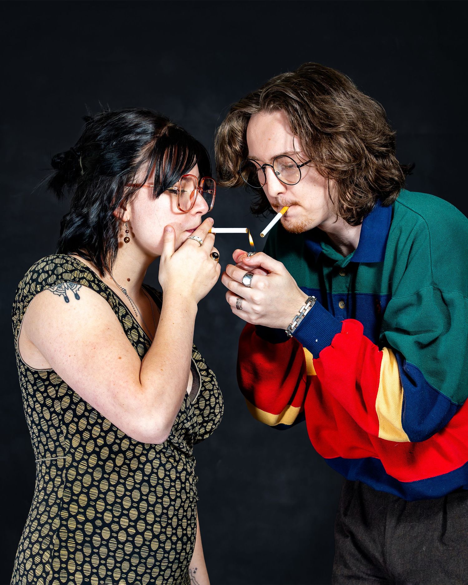 Two young people lighting cigarettes together against a dark background, wearing colorful retro clothing and glasses.