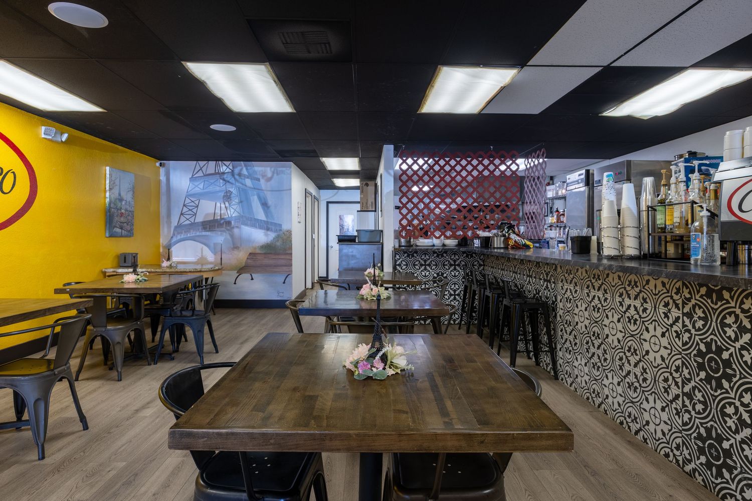 Modern restaurant interior featuring rustic wooden tables, decorative tile wall, and a bar area with black ceiling.