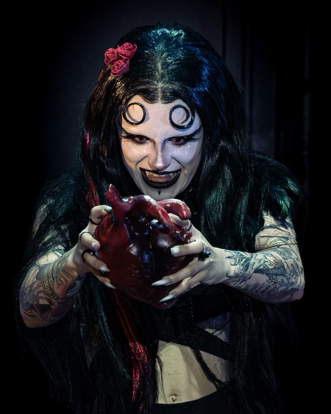 Tattooed performer in gothic vampire makeup holds prop heart wearing dark feathered costume with rose hair.