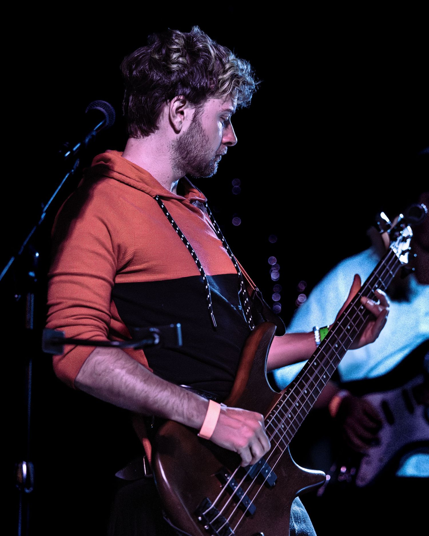 Bearded bassist in an orange shirt playing a brown bass guitar on a dark stage.