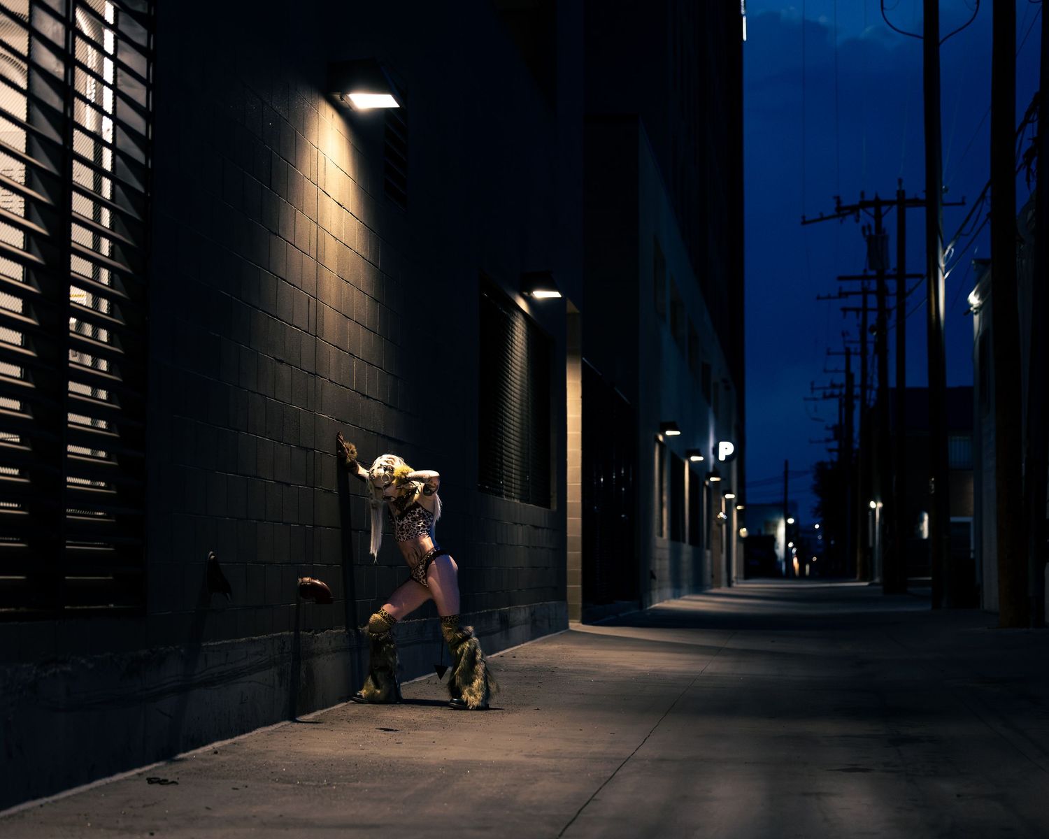 A lone figure leans against a dark alley wall at night, illuminated by street lights in an urban setting.