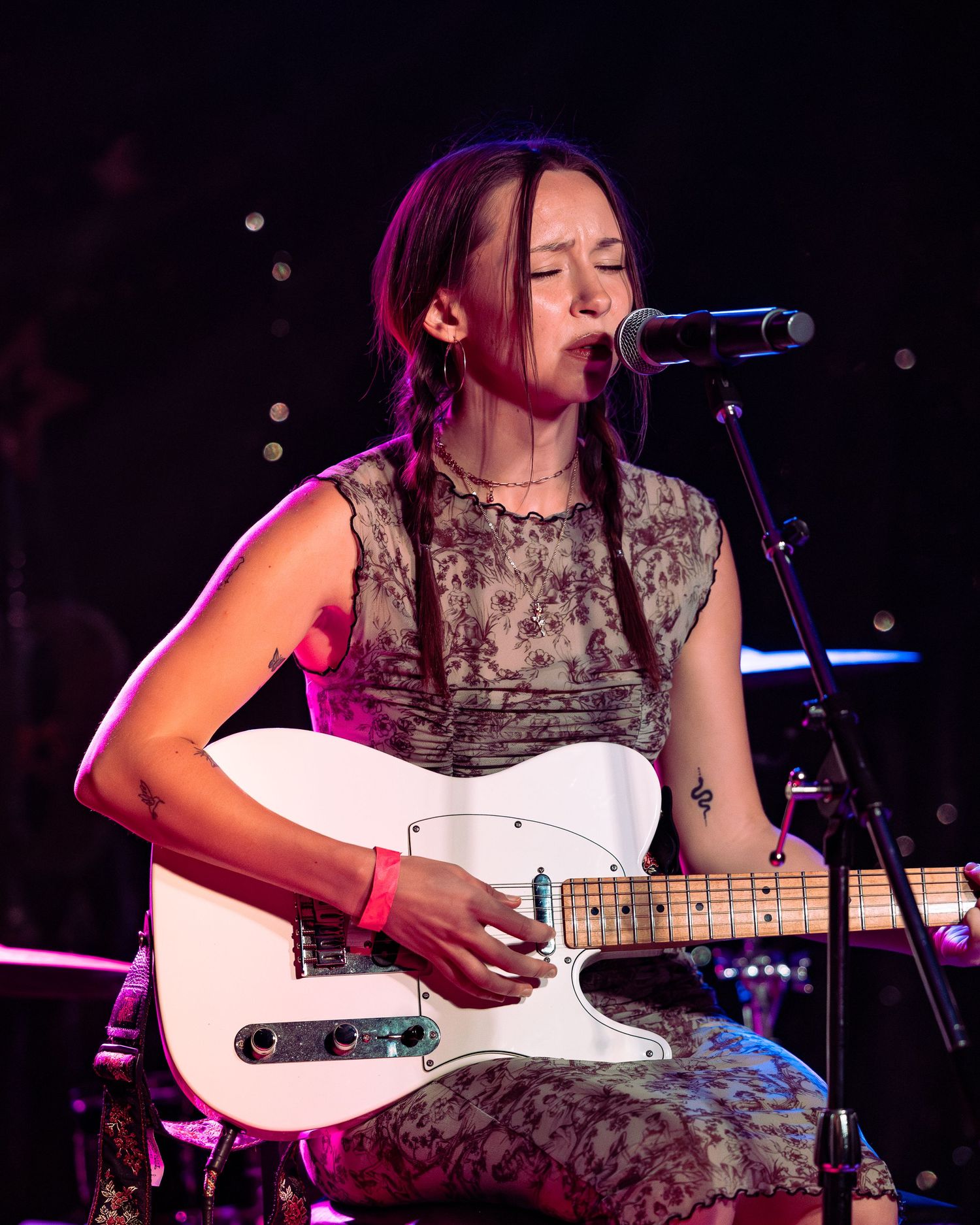 Young woman with braids playing white Telecaster guitar and singing into microphone on stage.