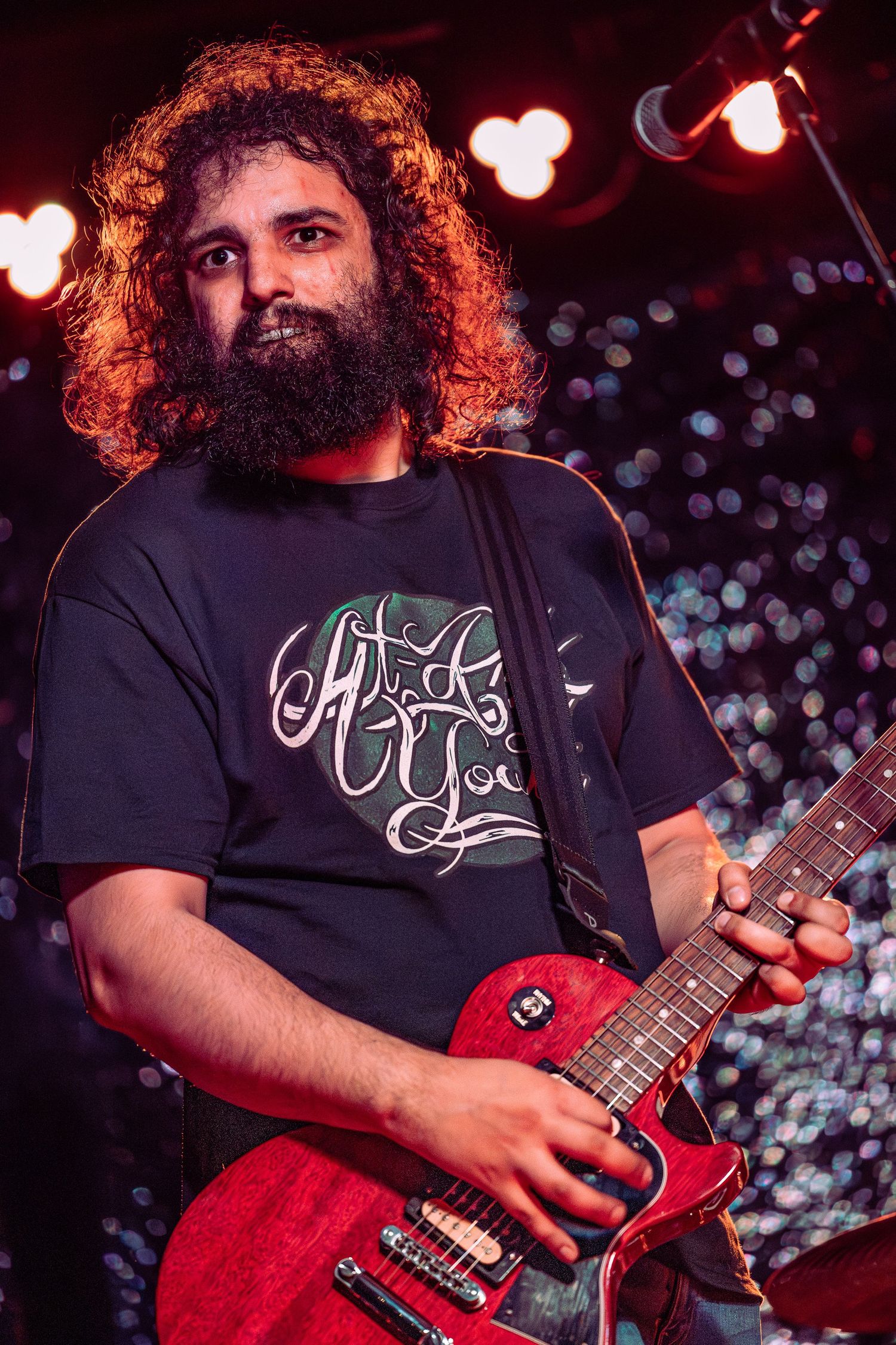 Bearded male guitarist with curly hair playing red electric guitar under warm orange stage lights.