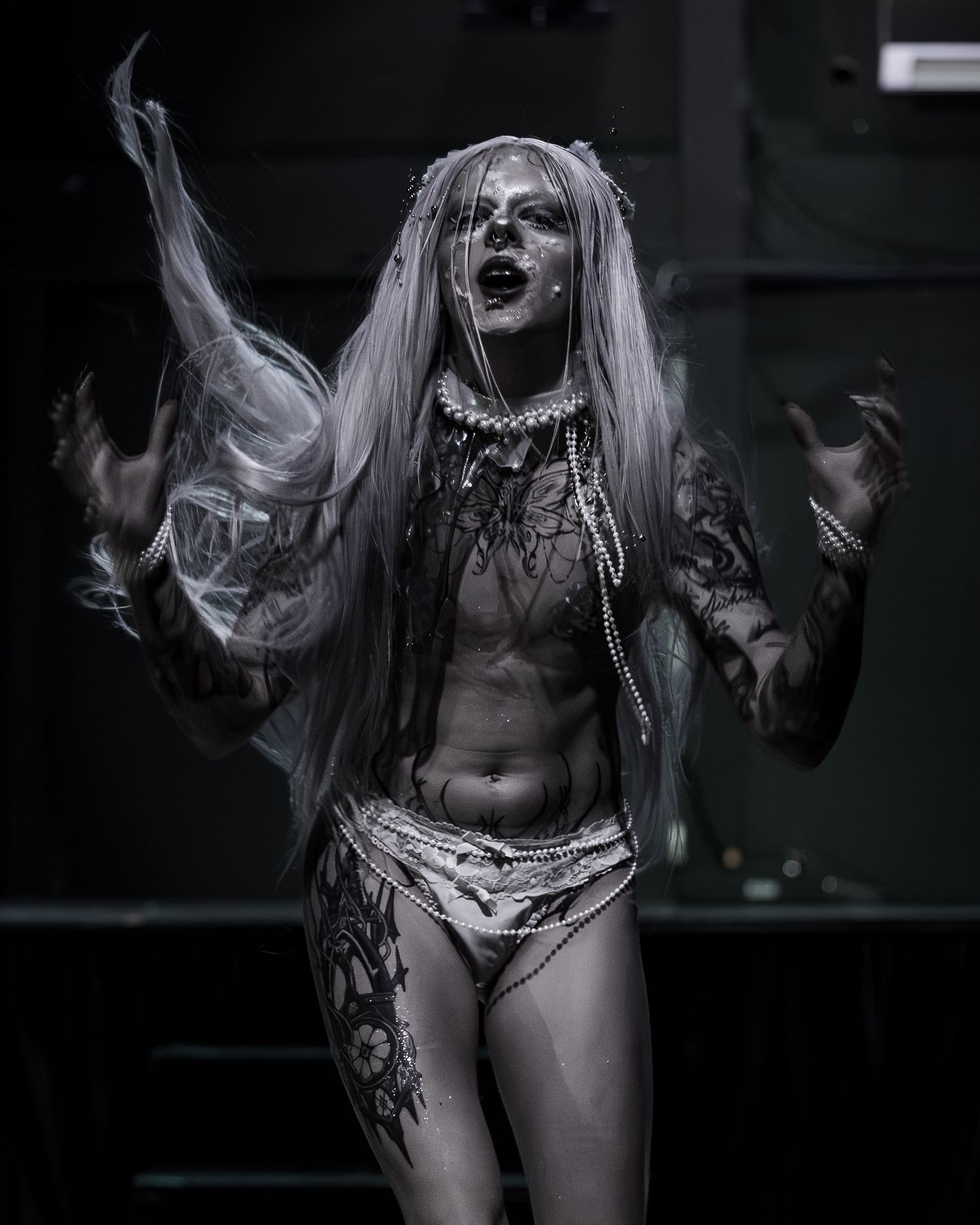 Heavily tattooed performer in gothic costume with white hair and dramatic makeup poses on stage in black and white photo.