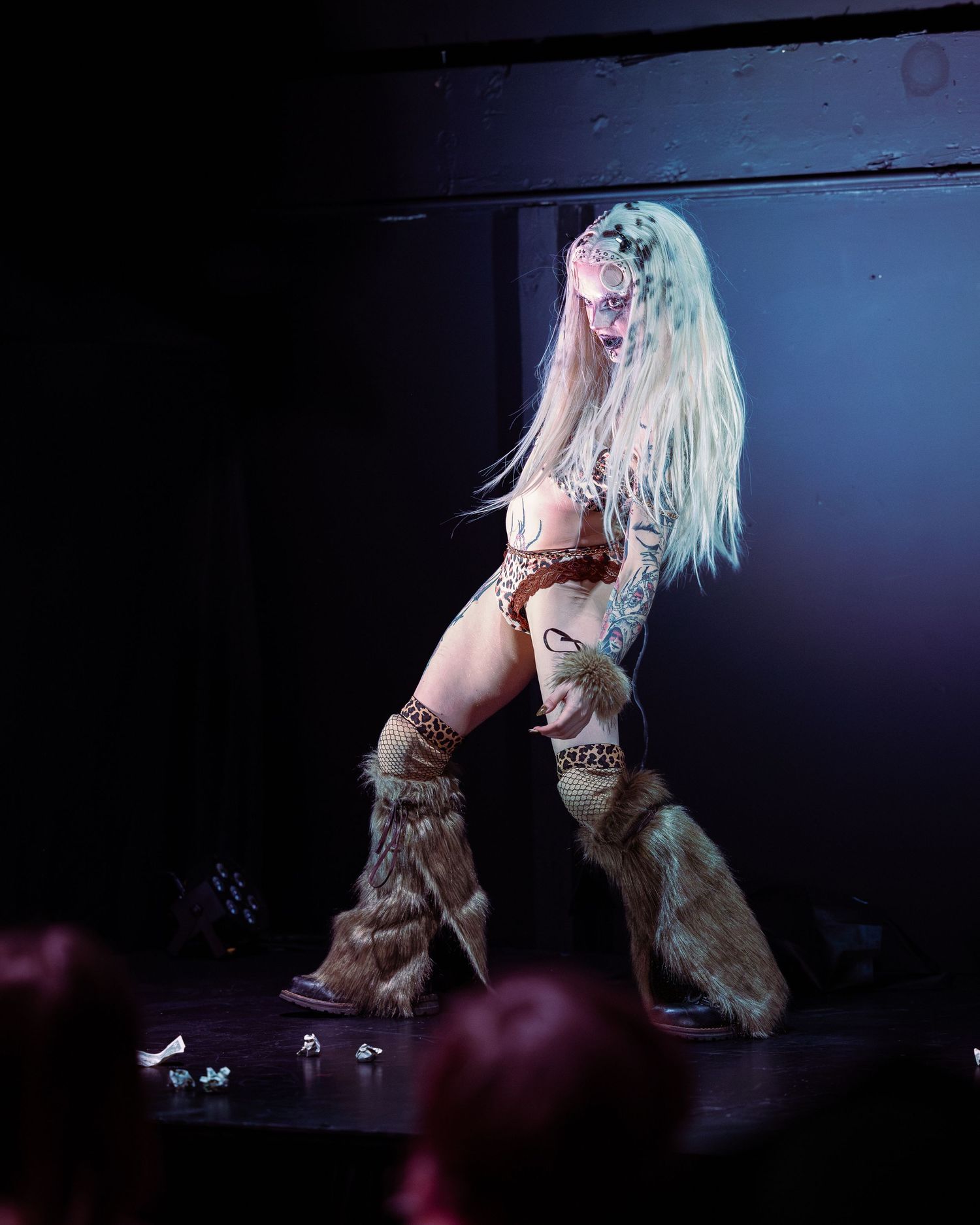 Tattooed burlesque dancer in leopard costume and fur boots performs on dark stage.