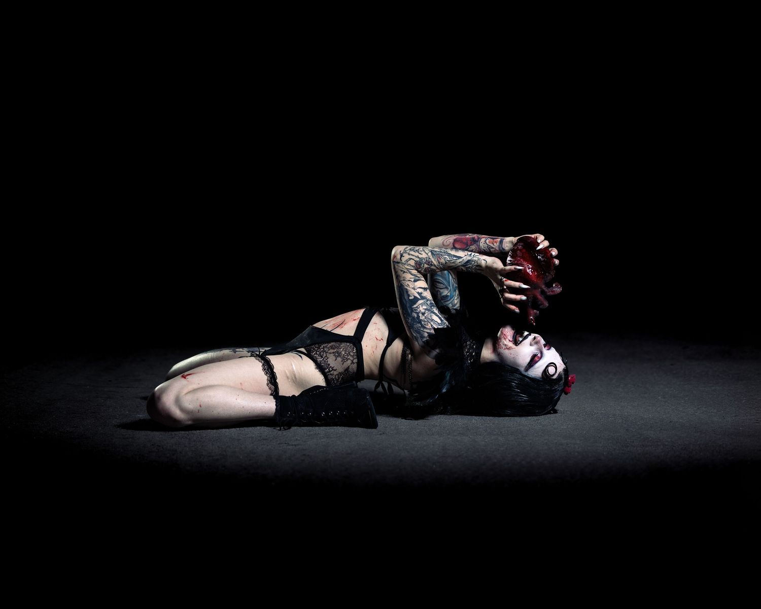 Two heavily tattooed performers in dramatic makeup lie entwined on a dark stage floor in artistic pose.