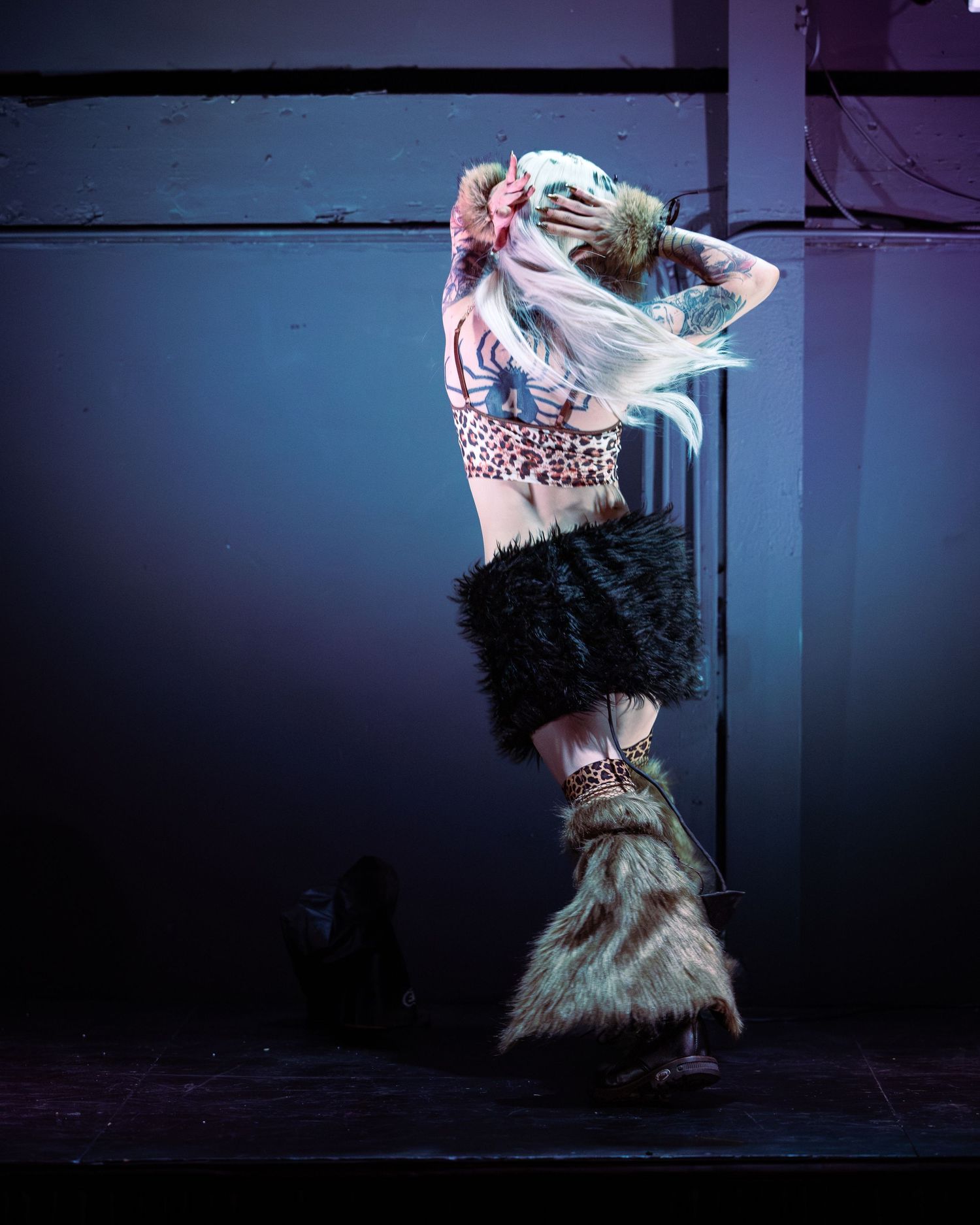 Performer in leopard crop top, black faux fur skirt, and fluffy leg warmers poses dramatically on stage.