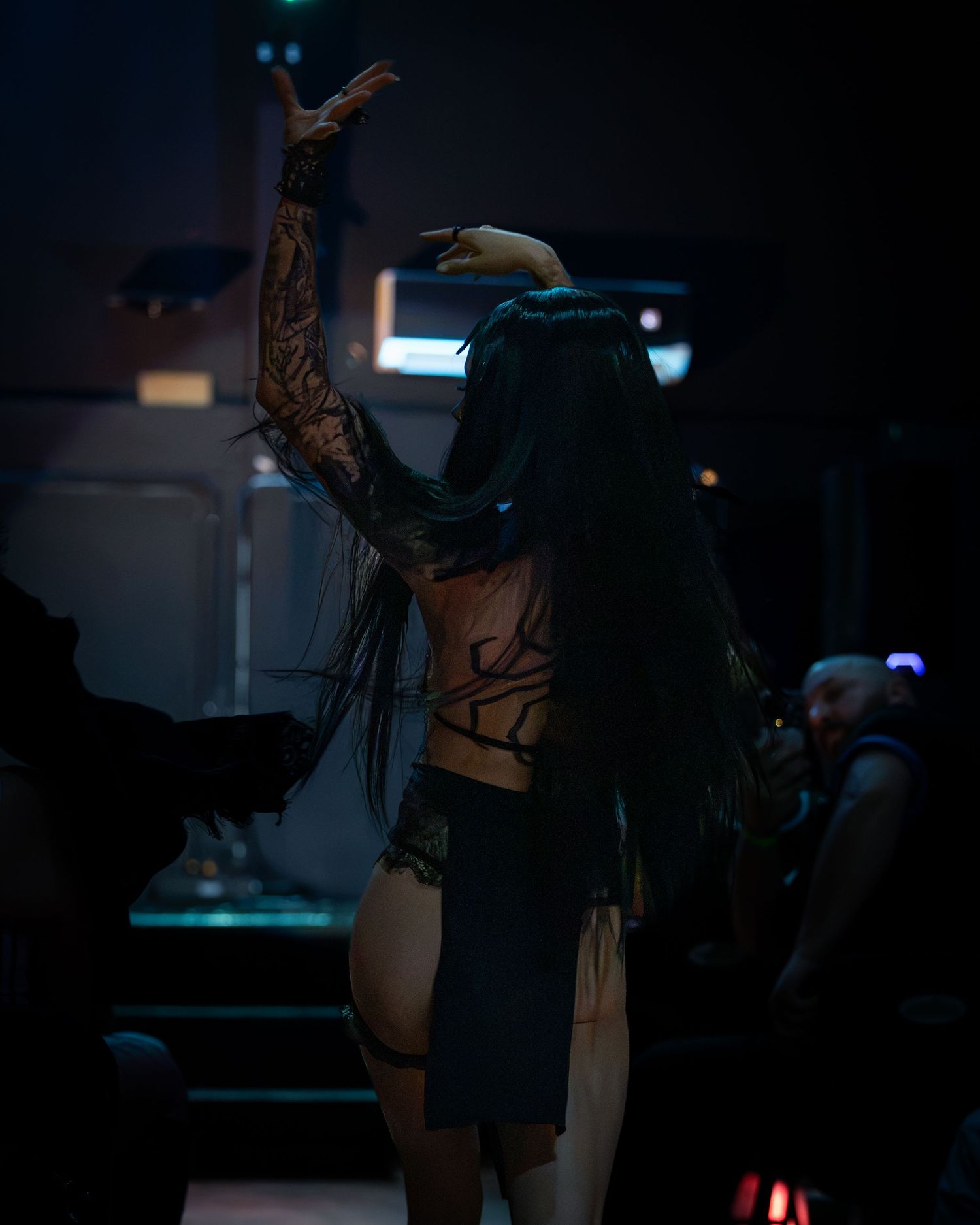 Tattooed performer with long dark hair raises arm on dimly lit stage during gothic performance.