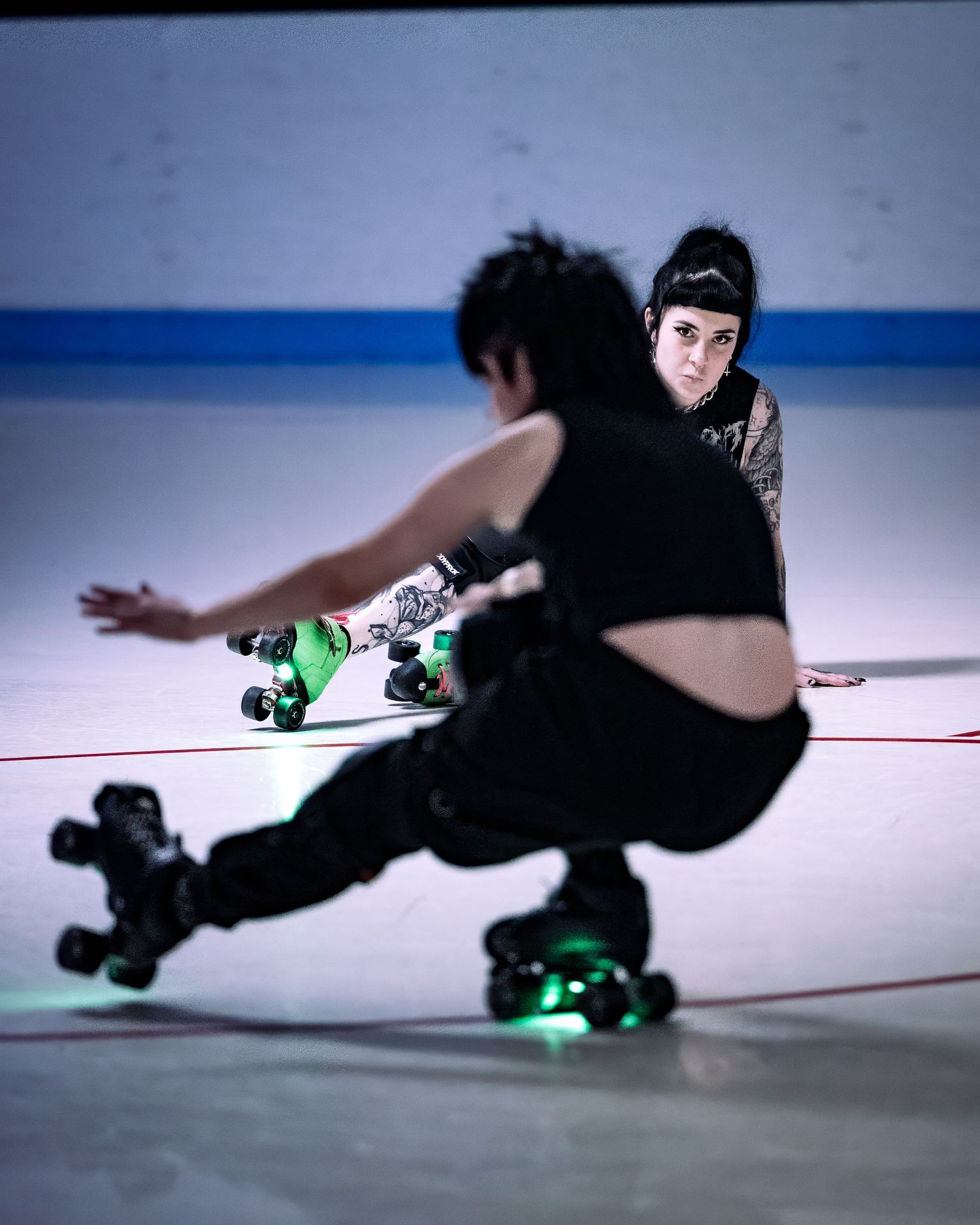 Two roller derby skaters in black gear compete on an indoor rink, wearing quad skates with green wheels.