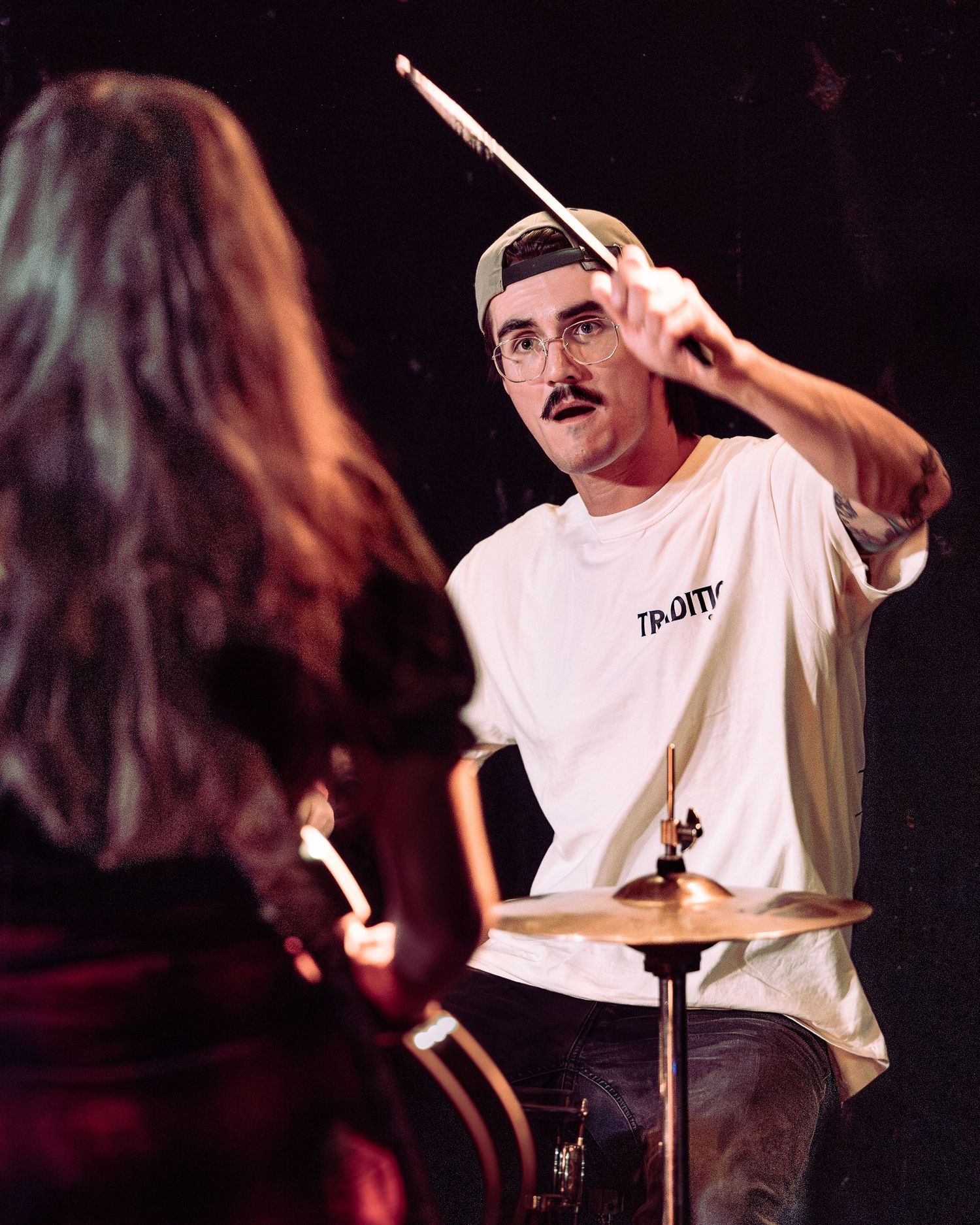 Drummer with mustache and cap raises drumstick over cymbal during live concert performance.