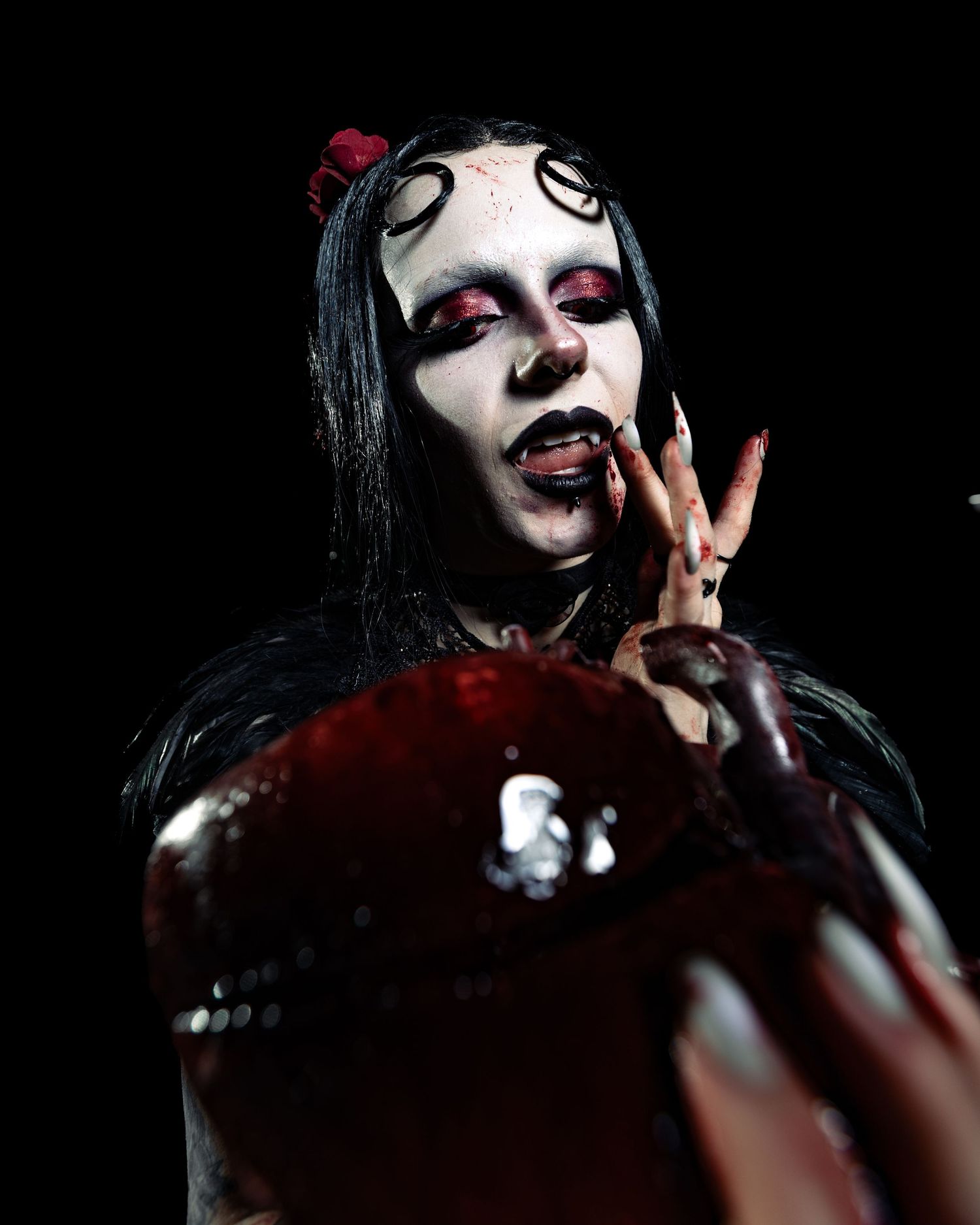 Gothic horror character with white face paint, spiral eye makeup, and blood effects holding red object.