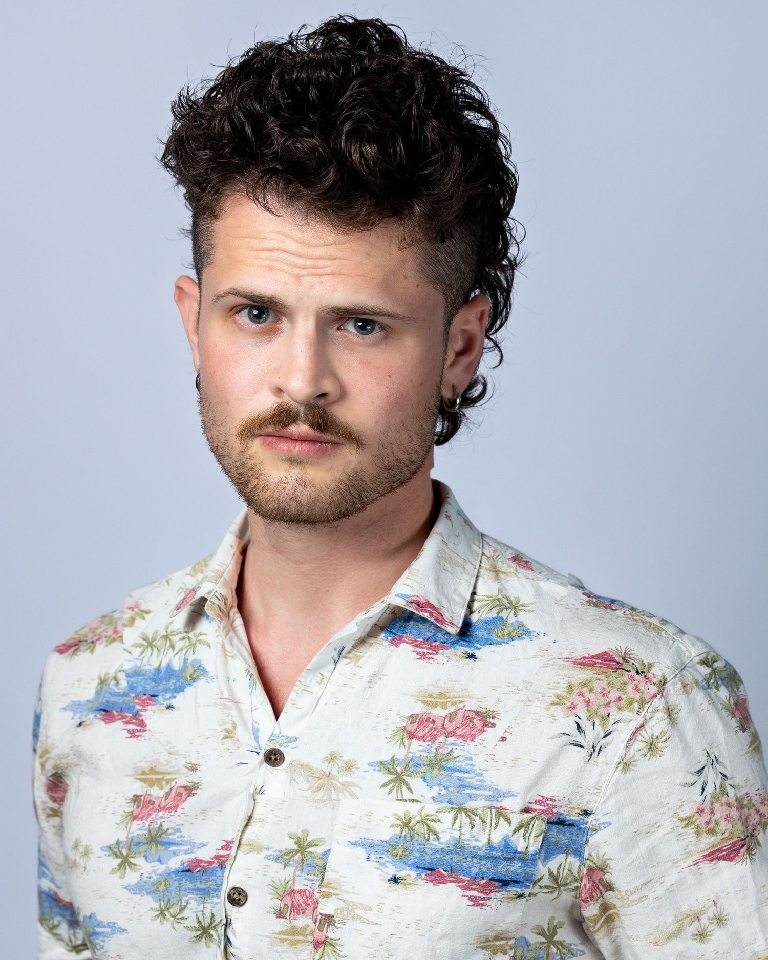 Young man with curly hair and mustache wearing a floral Hawaiian shirt poses for a professional headshot.