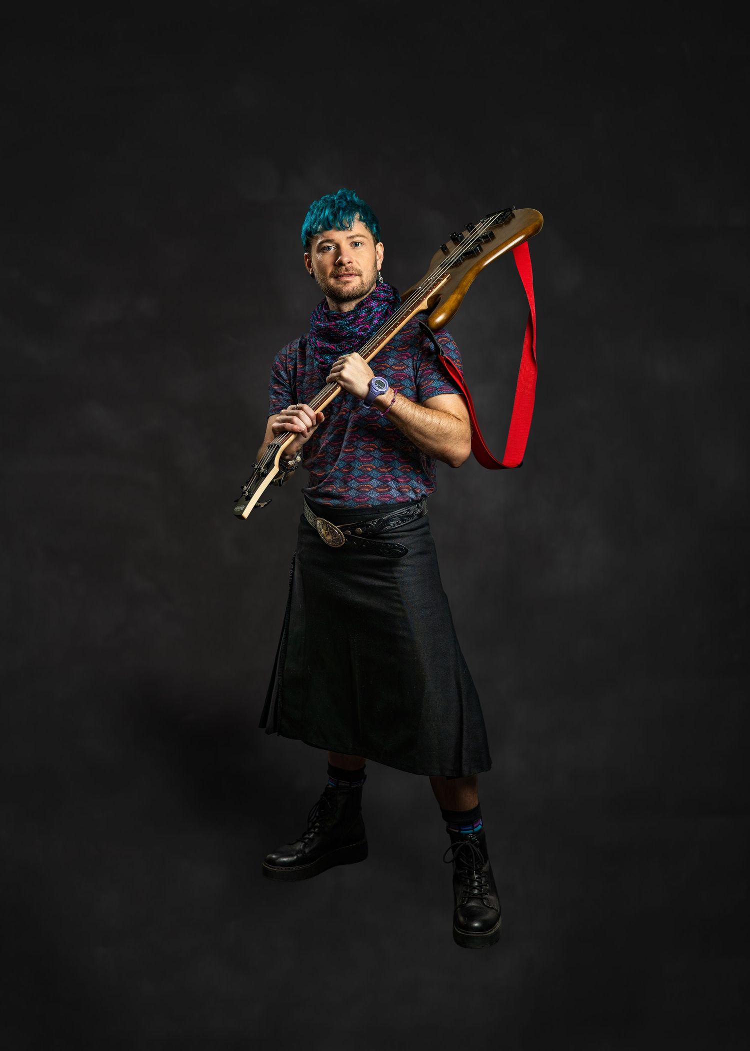 Musician with teal hair in a black kilt plays a gold flute with a red strap against a dark background.