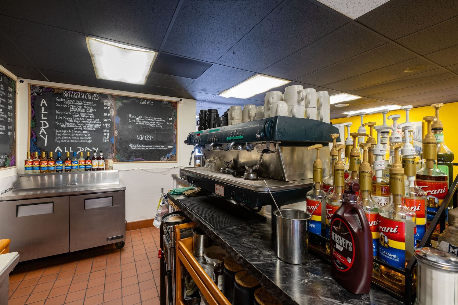 Commercial espresso machine and bar area with syrups, chalkboard menu, and refrigerated counter in a cafe setting.