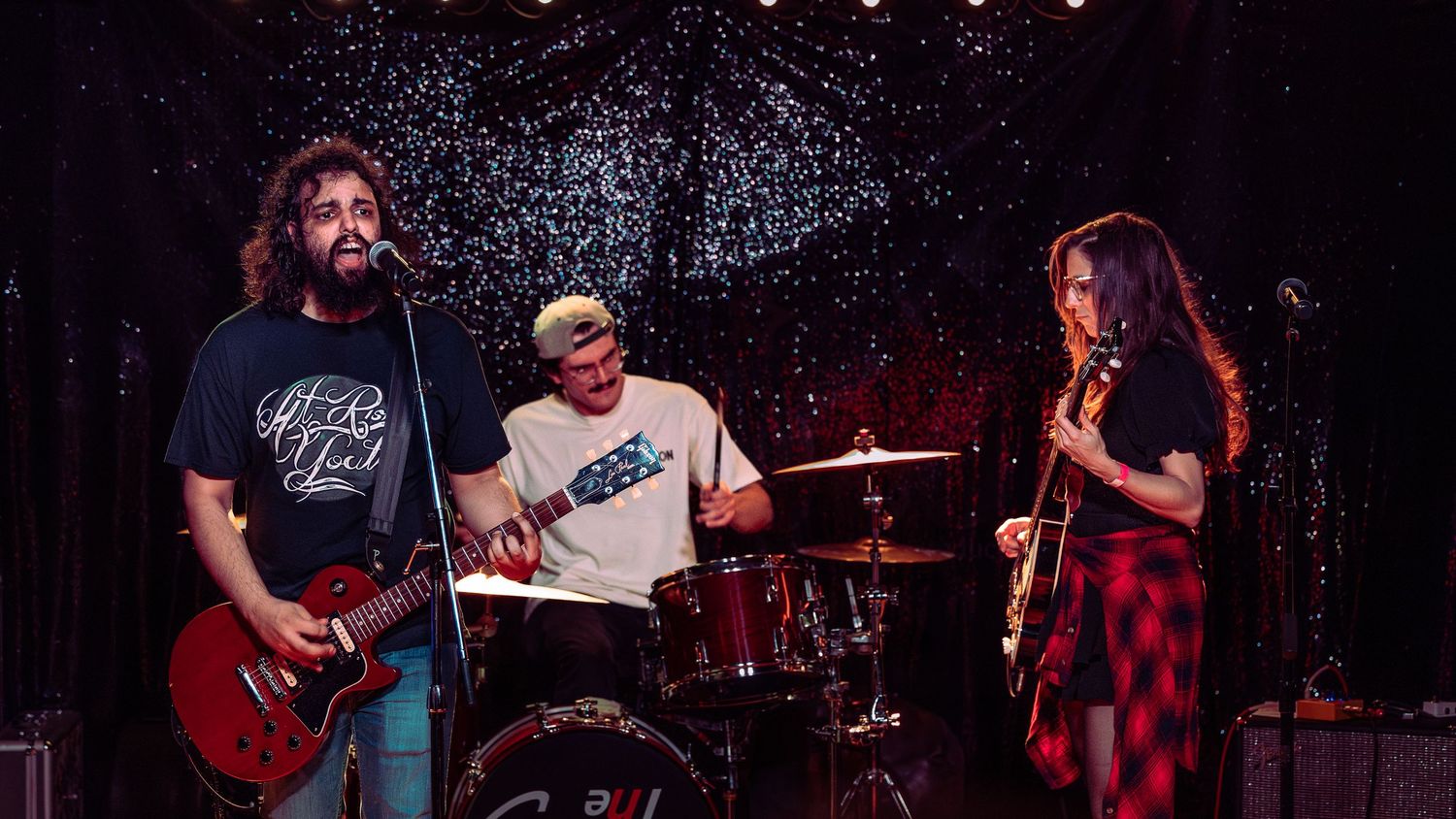 Three-piece band performing on stage with guitarist, drummer, and female guitarist under warm stage lights.