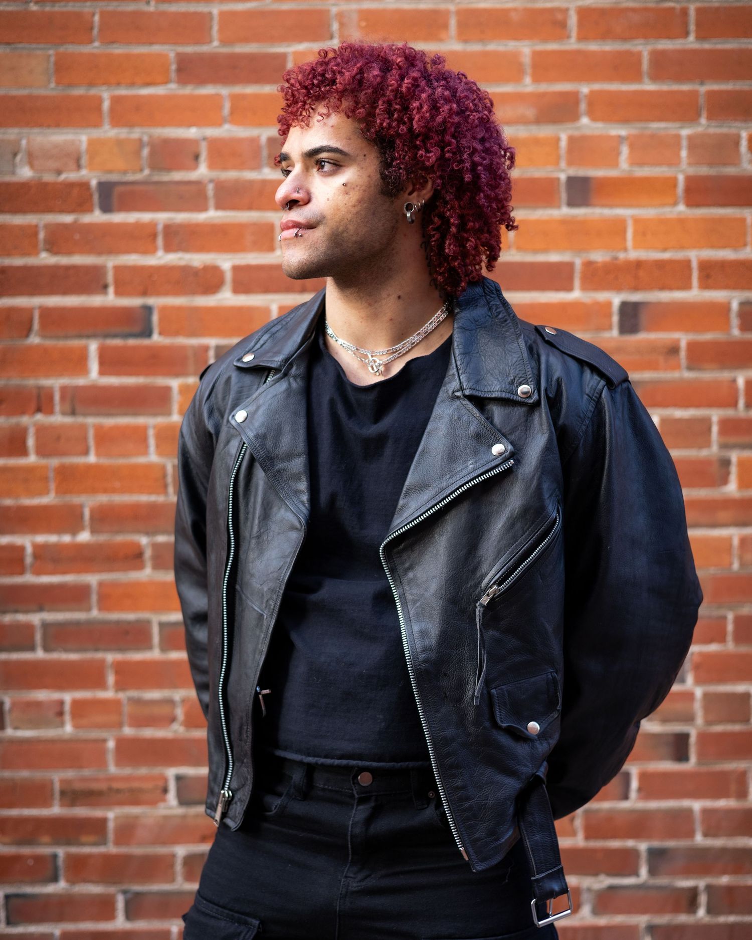 Stylish leather jacket and magenta curly hair look against rustic brick wall background.
