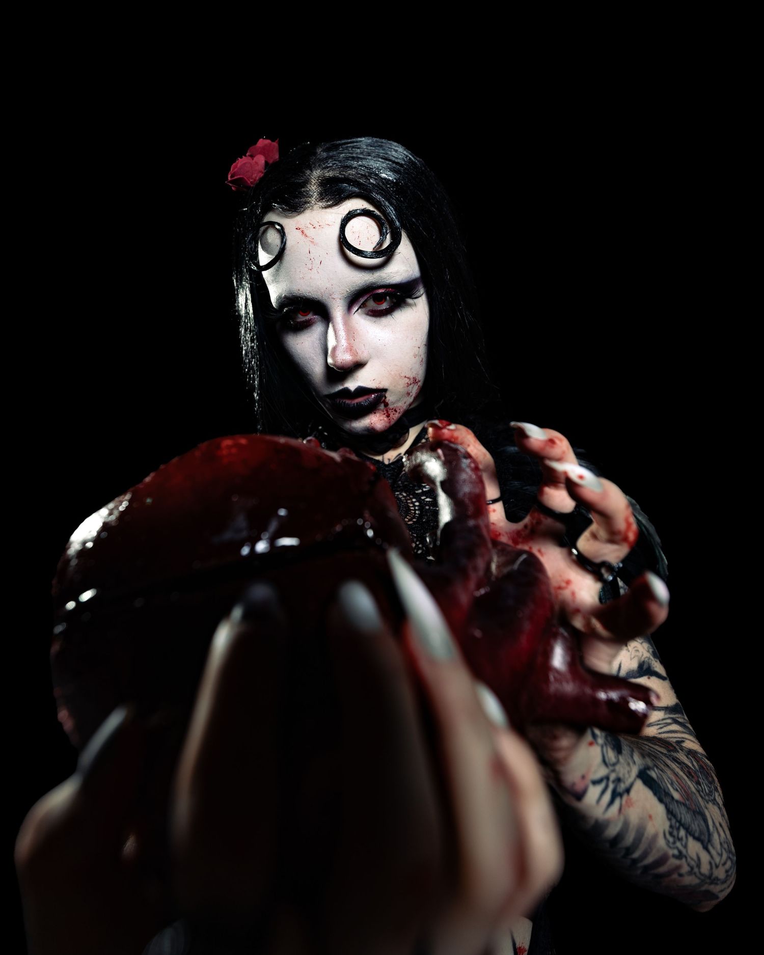 Gothic woman with spiral eye makeup, blood-covered hands holding a heart, dark Halloween costume with tattoos.