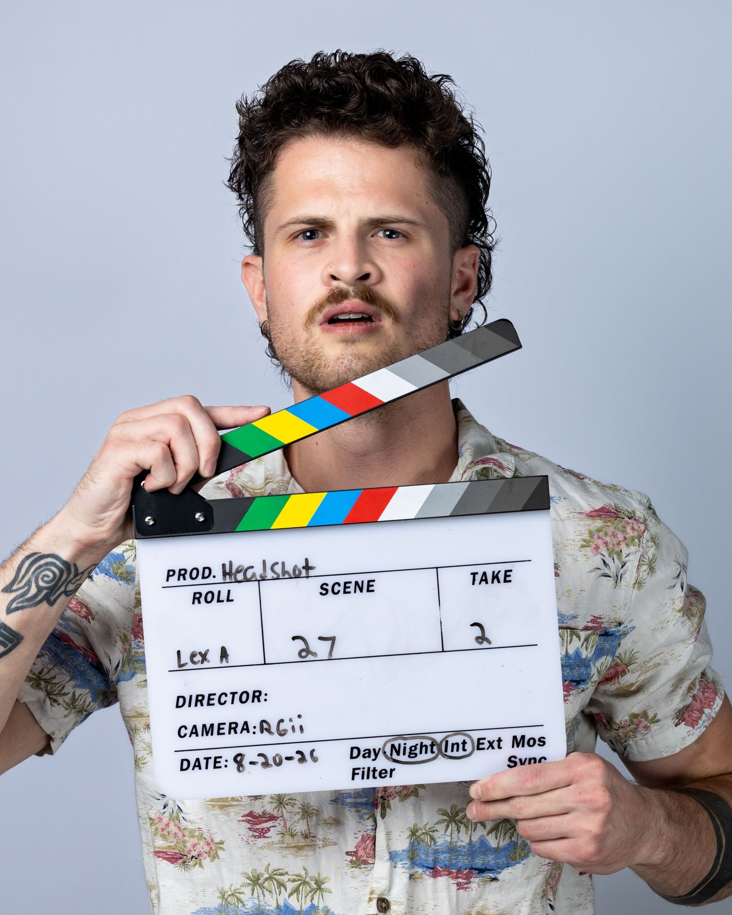 Young man with curly hair holding a film clapperboard, wearing a floral shirt, on a studio set.