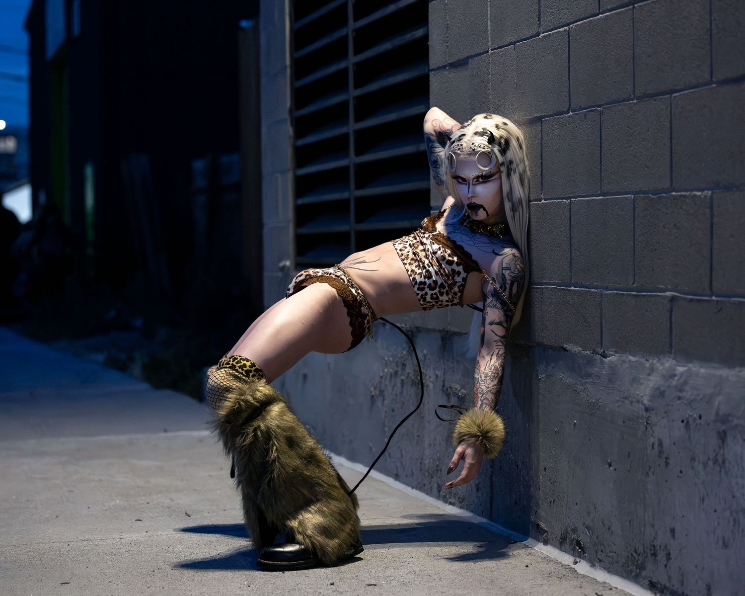 Tattooed woman in leopard print costume with fur boots poses against urban wall at night.