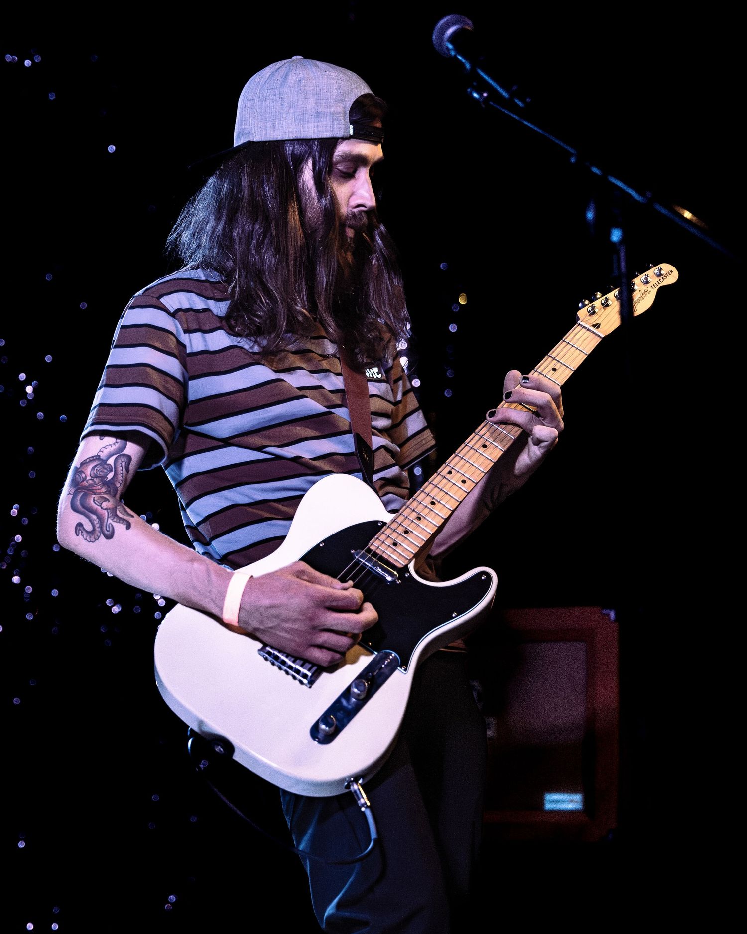 Long-haired guitarist in striped shirt plays white Telecaster guitar on stage.