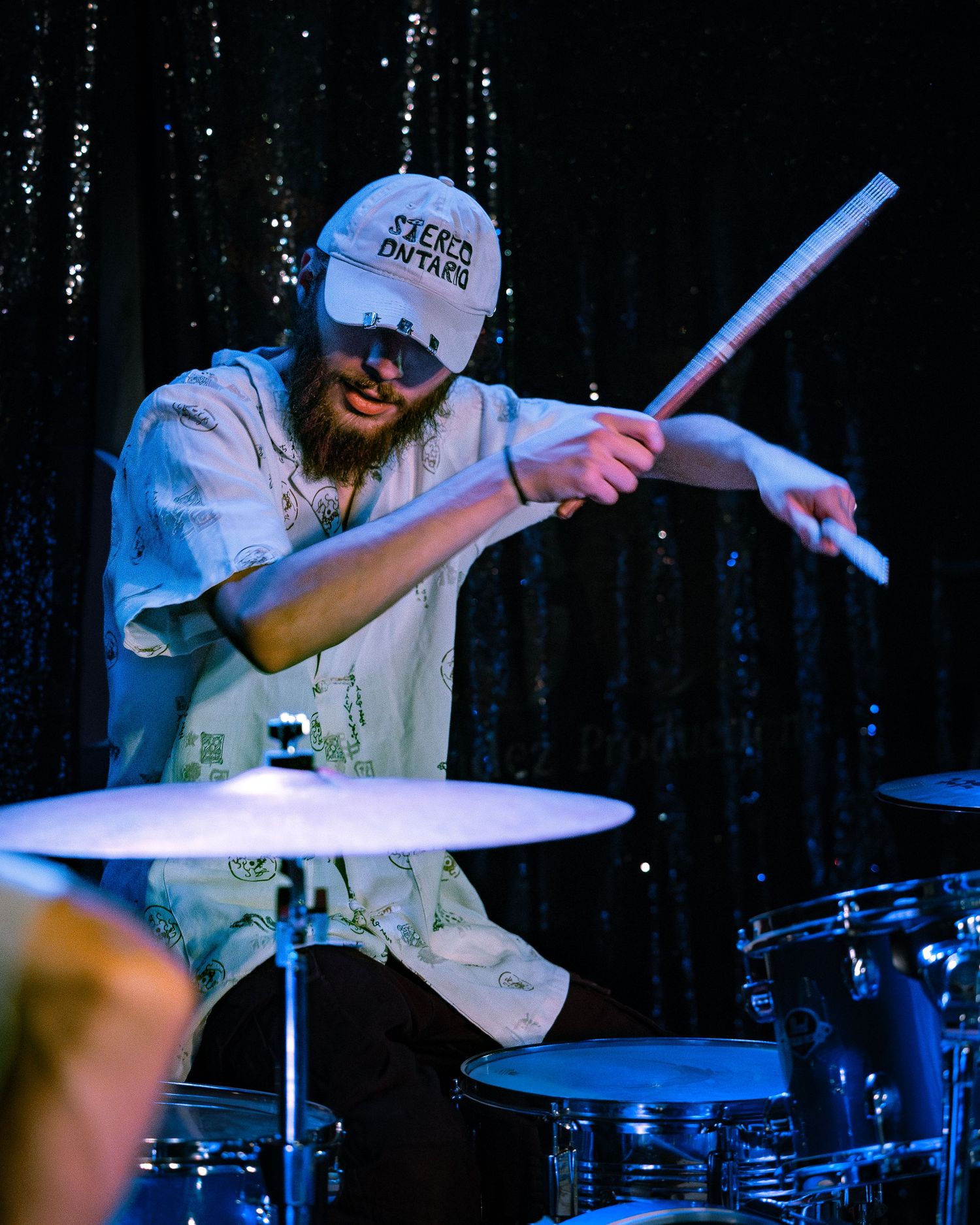 Bearded drummer in white cap energetically plays cymbals with drumsticks under blue concert lighting.