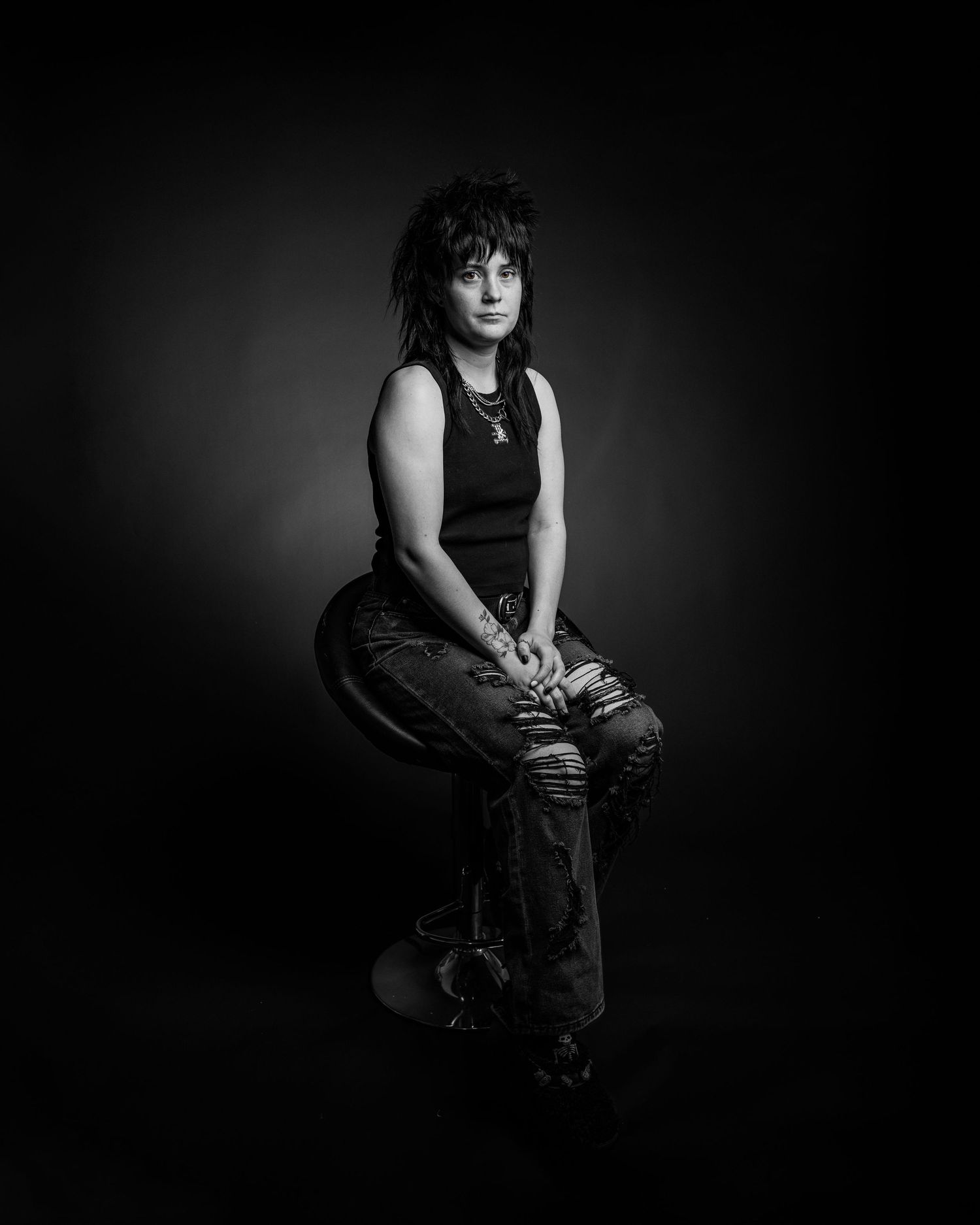 Black and white portrait of a person with dark mullet hair sitting on a stool in a sleeveless top and jeans.