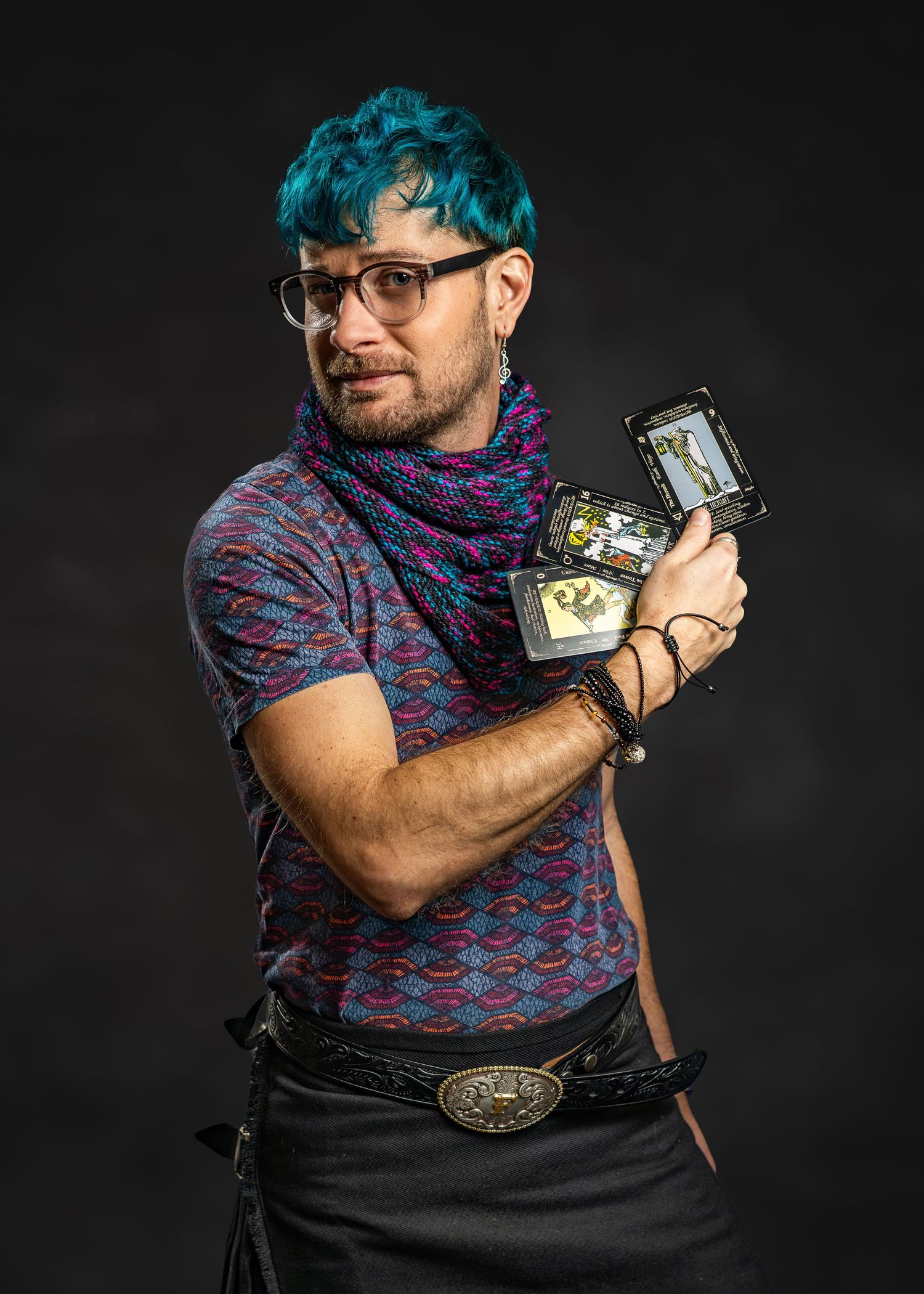 Person with teal hair and glasses holding trading cards, wearing colorful scarf and patterned shirt against dark background.