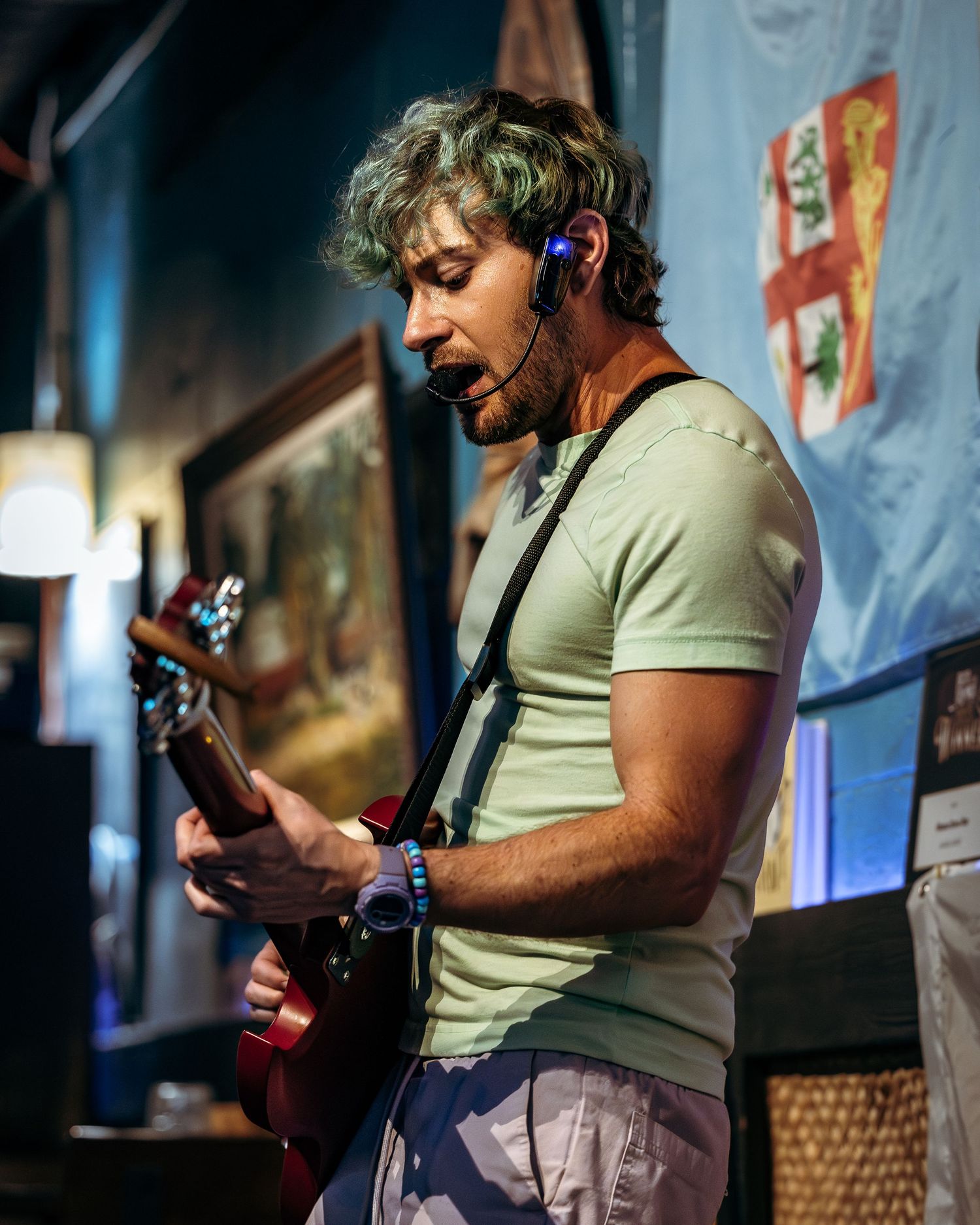Musician performs on stage with a guitar and headset microphone while wearing a light-colored t-shirt.