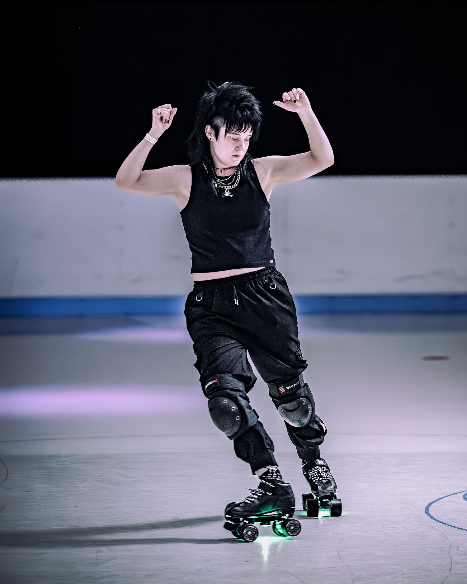 Person roller skating in black outfit with knee pads, striking a dynamic pose on an indoor rink.