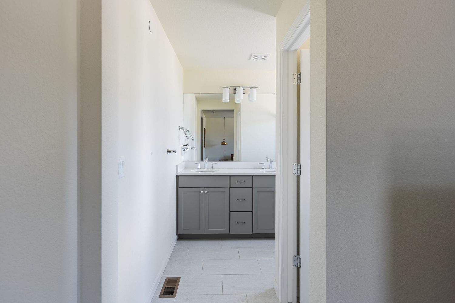Hallway view leading to a modern bathroom featuring gray cabinets, white countertop, and large vanity mirror.
