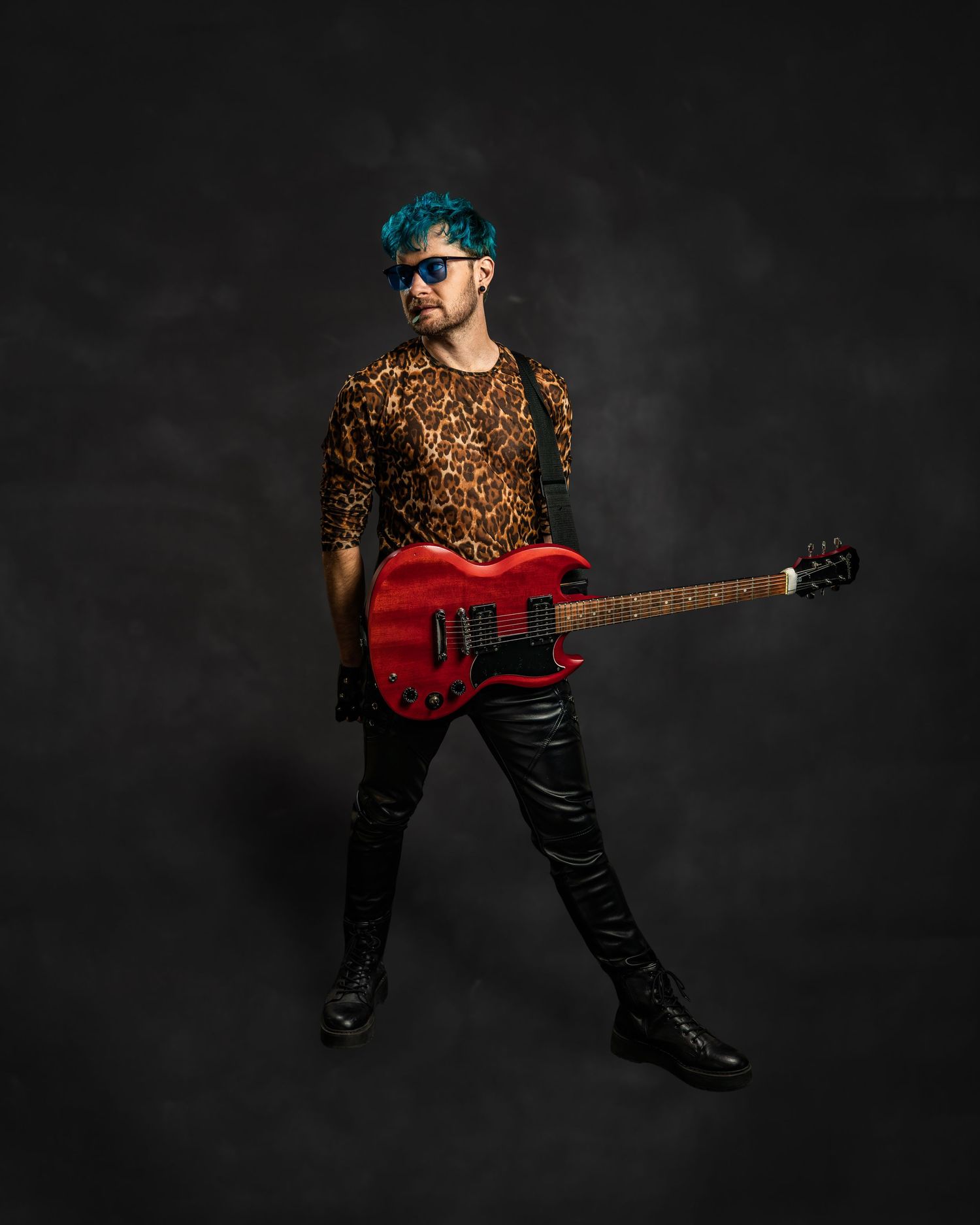 Rock musician with teal hair in leopard print shirt holds red electric guitar wearing black leather pants.