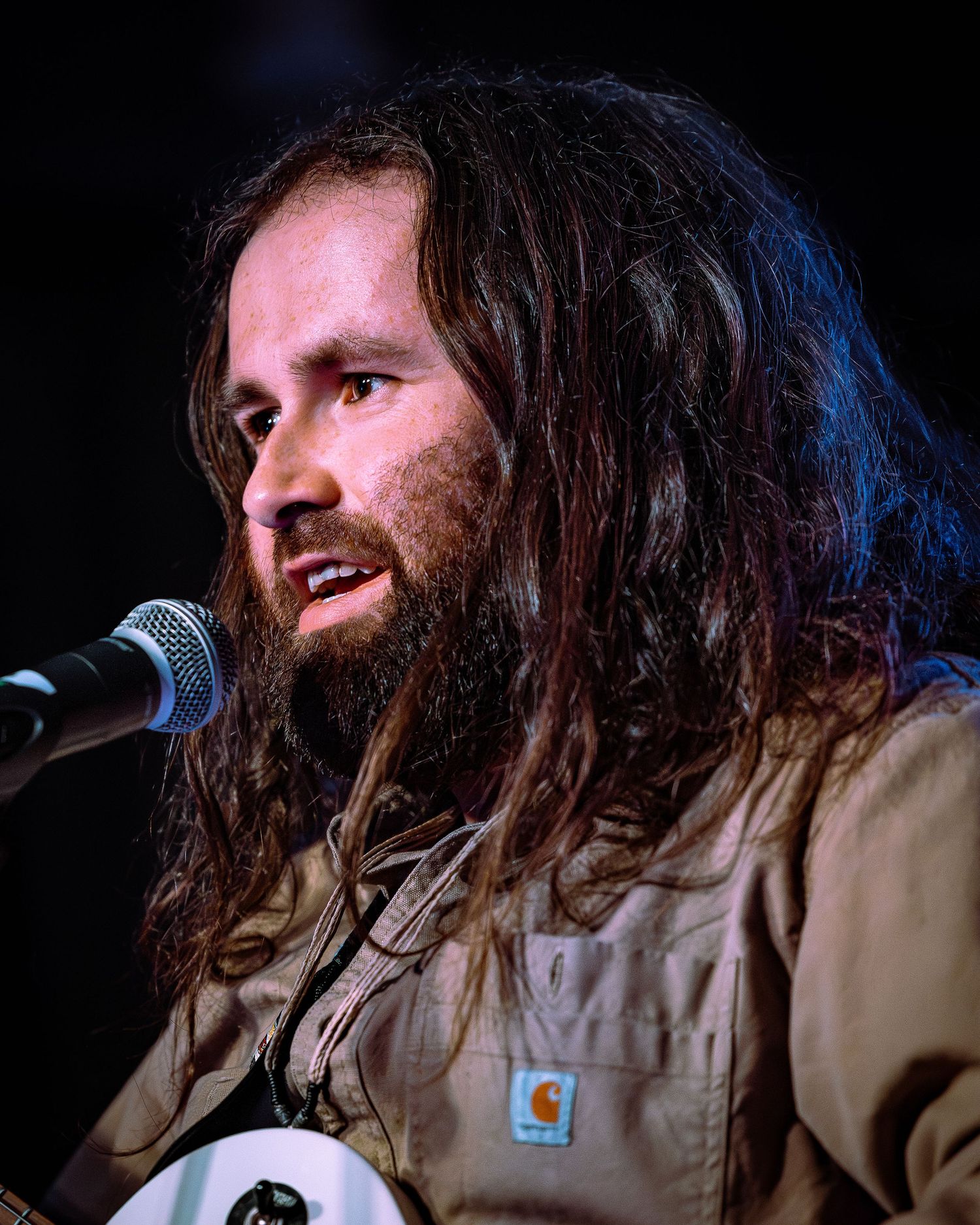 Long-haired bearded male musician speaks into microphone wearing a tan Carhartt jacket at live event.