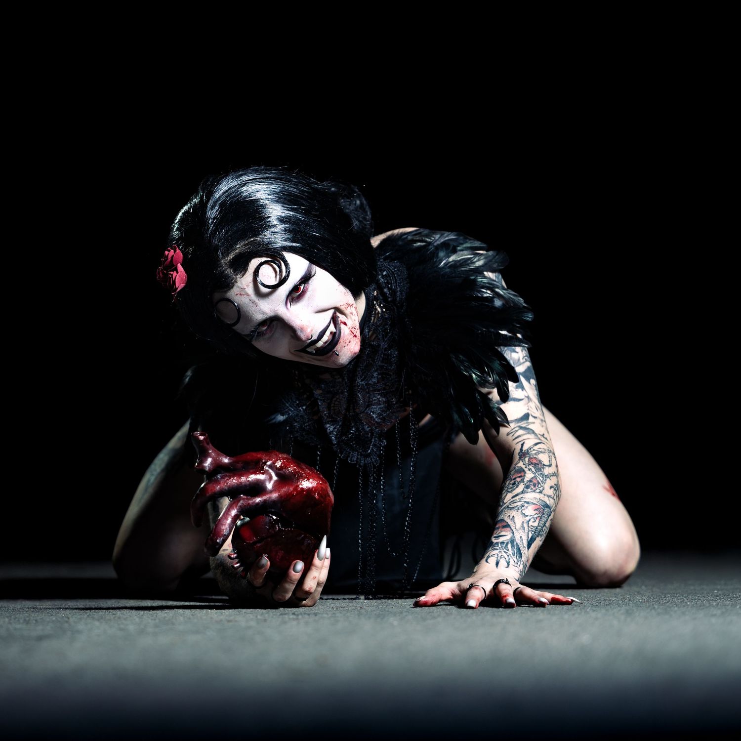 Woman in dramatic Halloween makeup with black feathers, tattoos, and fake blood in dark studio setting.