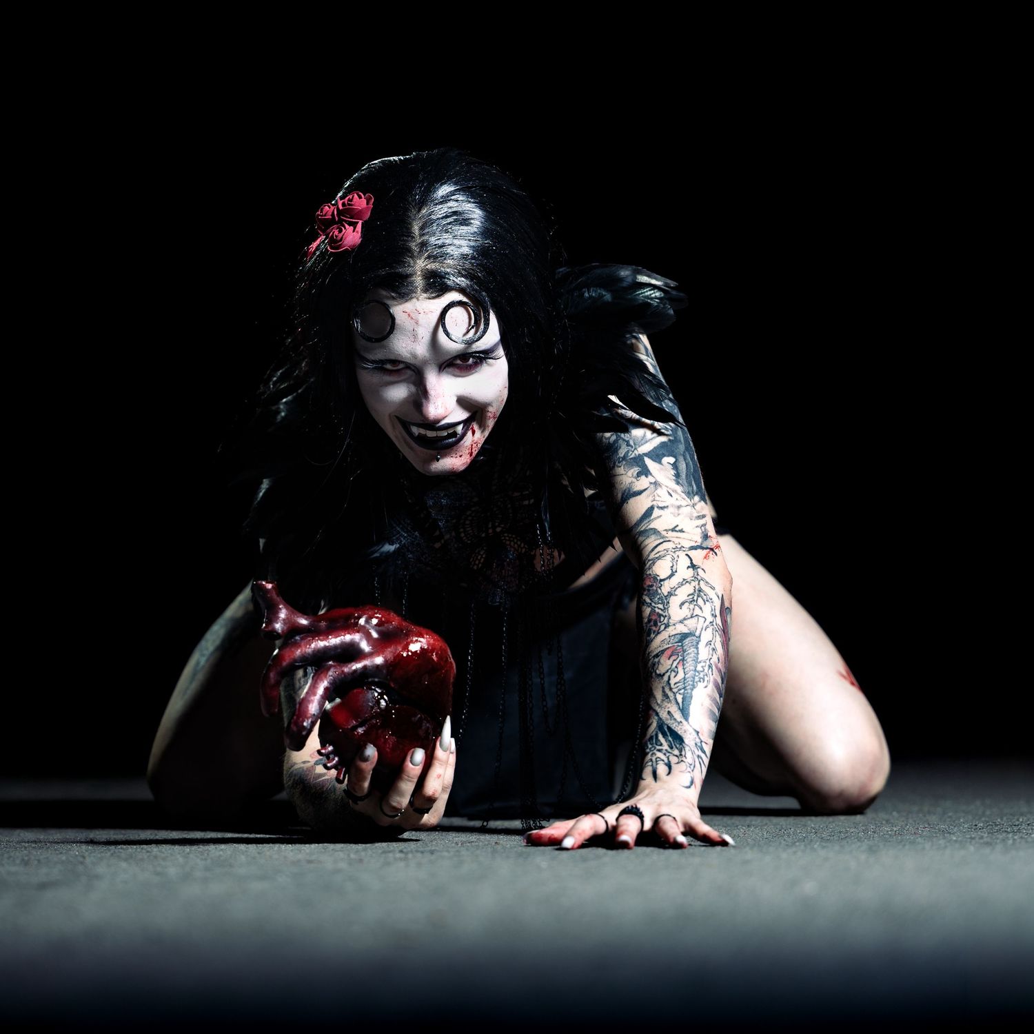 Tattooed woman in zombie Halloween costume with dark makeup crawling on floor holding bloody heart prop.