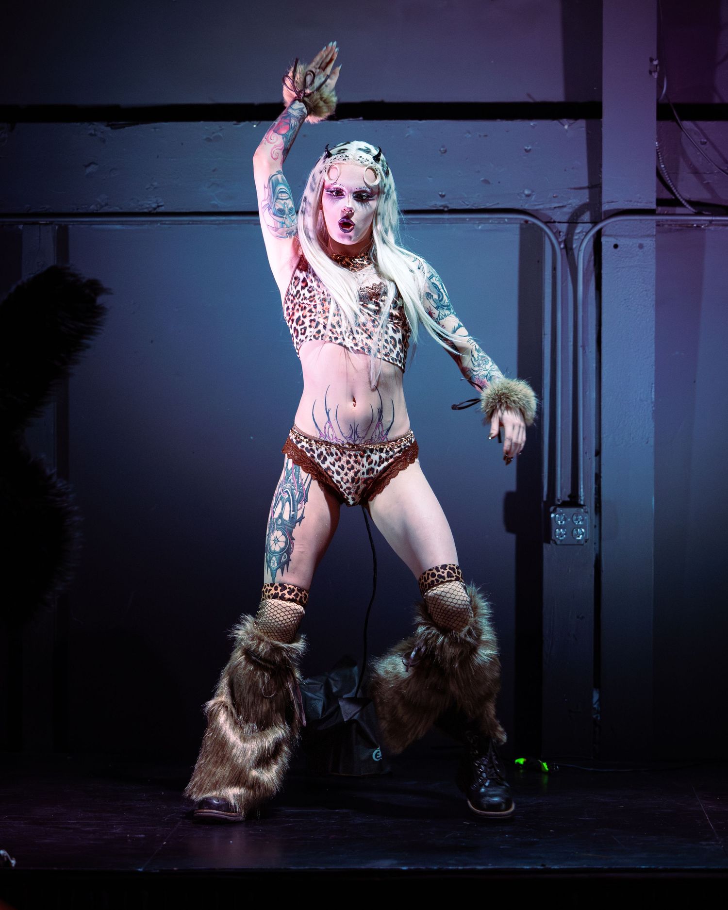 Tattooed performer in leopard print outfit and fur boots poses dramatically on stage under purple lighting.