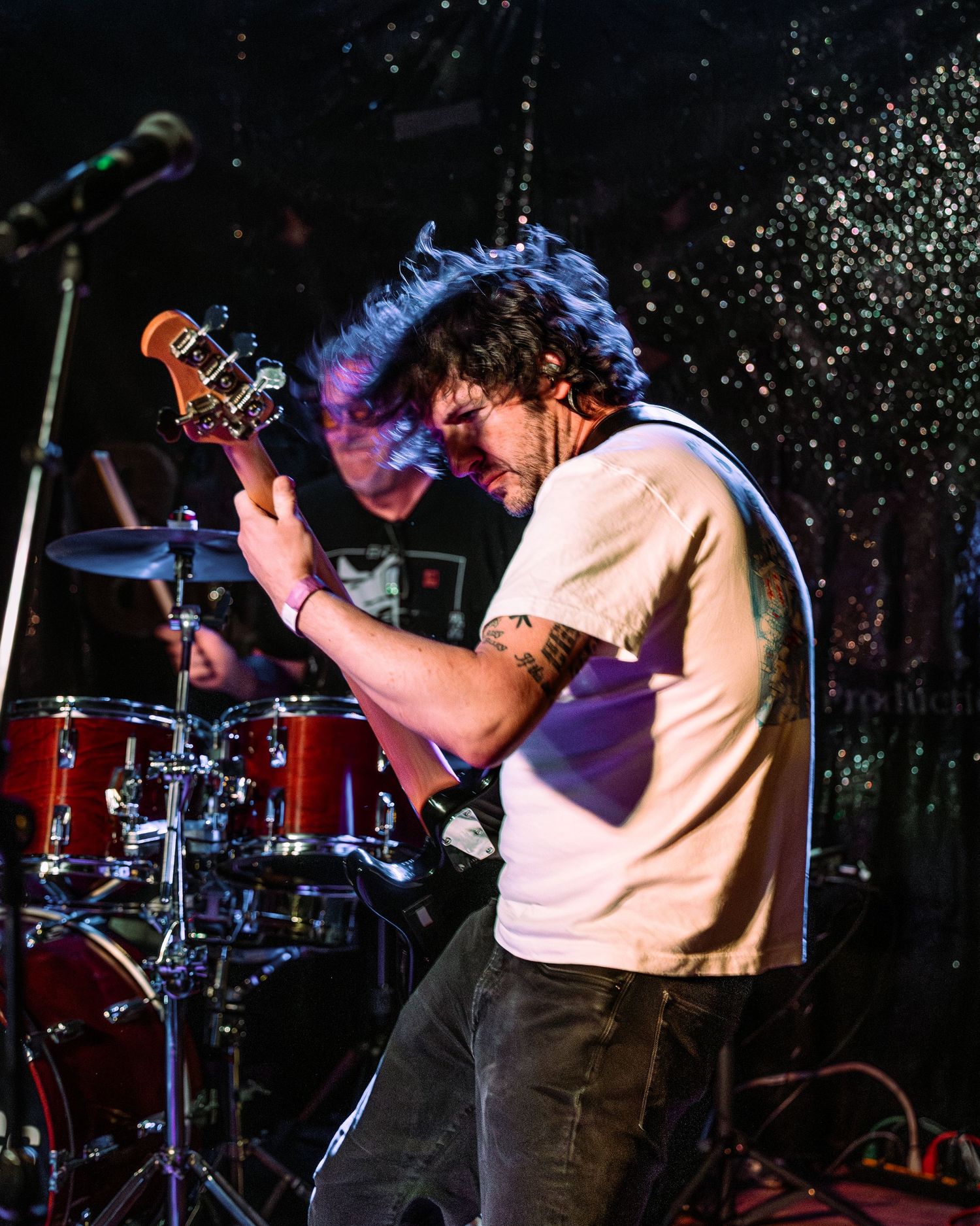 Energetic guitarist headbanging on stage with tattoos visible, drummer visible in background.