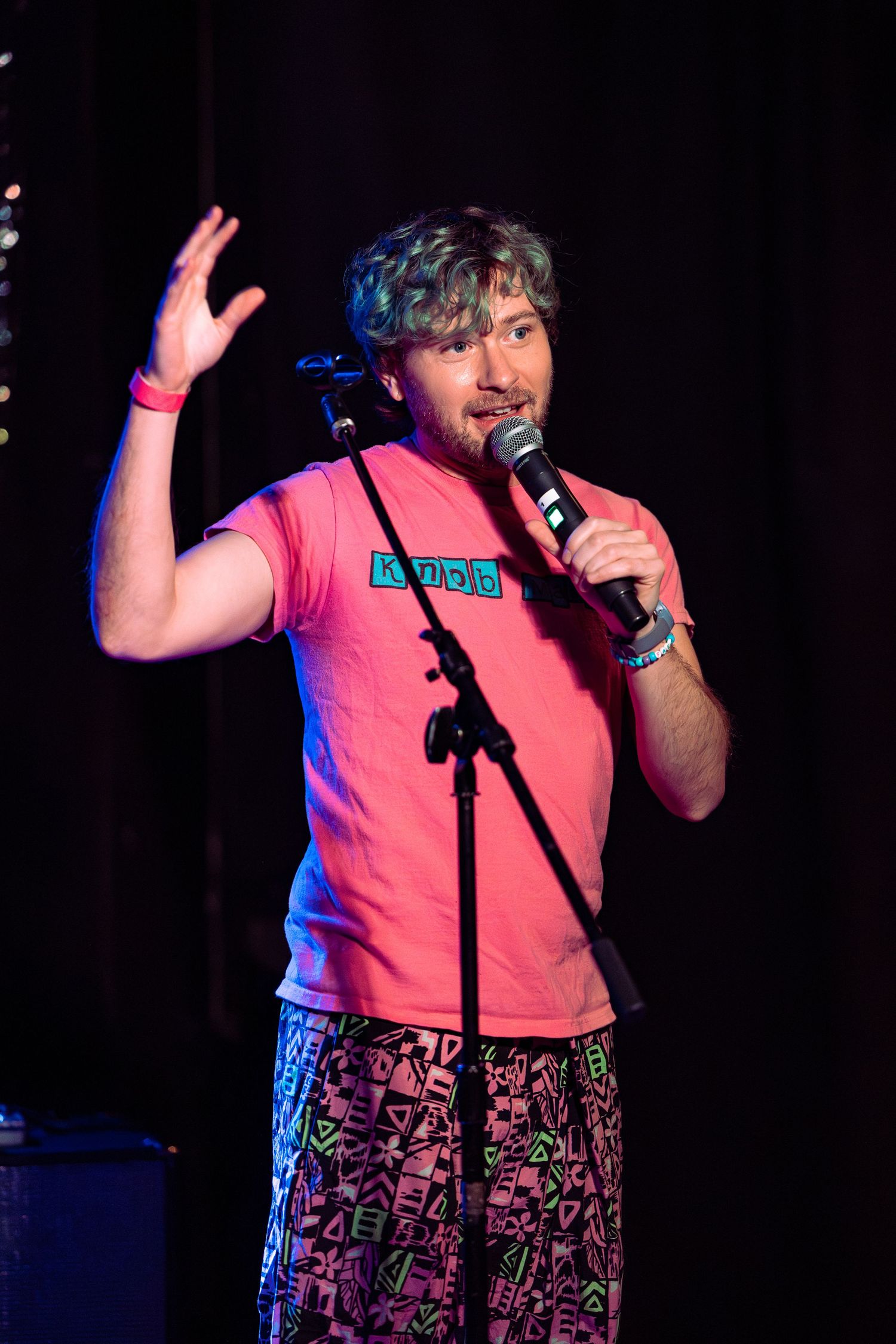 Man in pink t-shirt and patterned shorts performs stand-up comedy on stage holding a microphone.