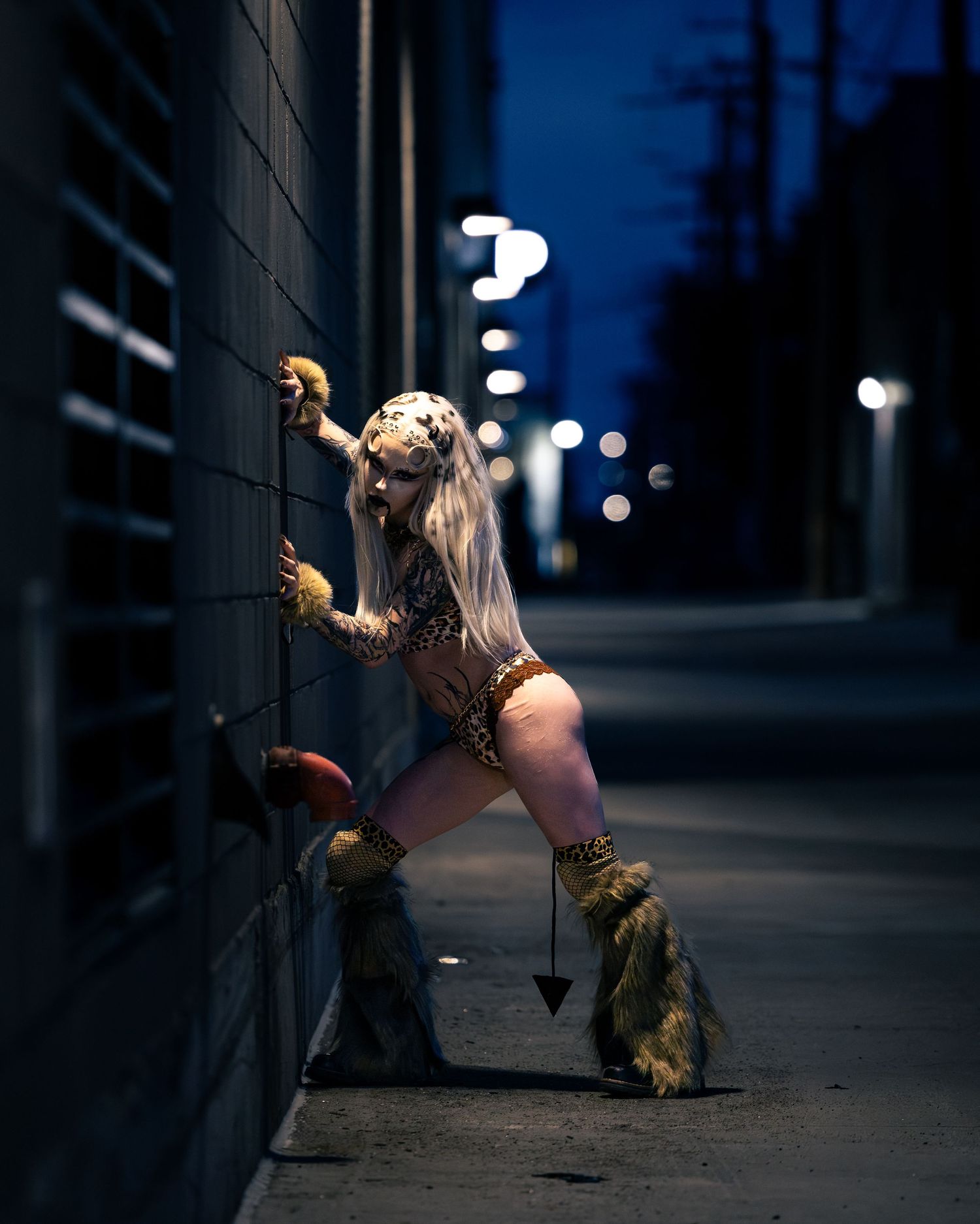 Woman in leopard costume and fur boots poses against alley wall at night in dramatic blue lighting.