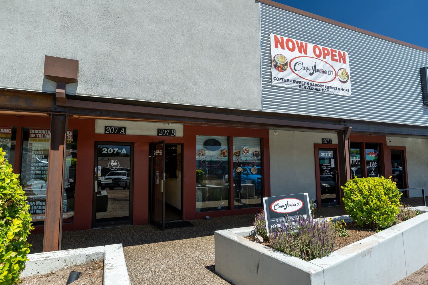 Exterior of a strip mall restaurant with a Now Open sign and outdoor seating area on a sunny day.